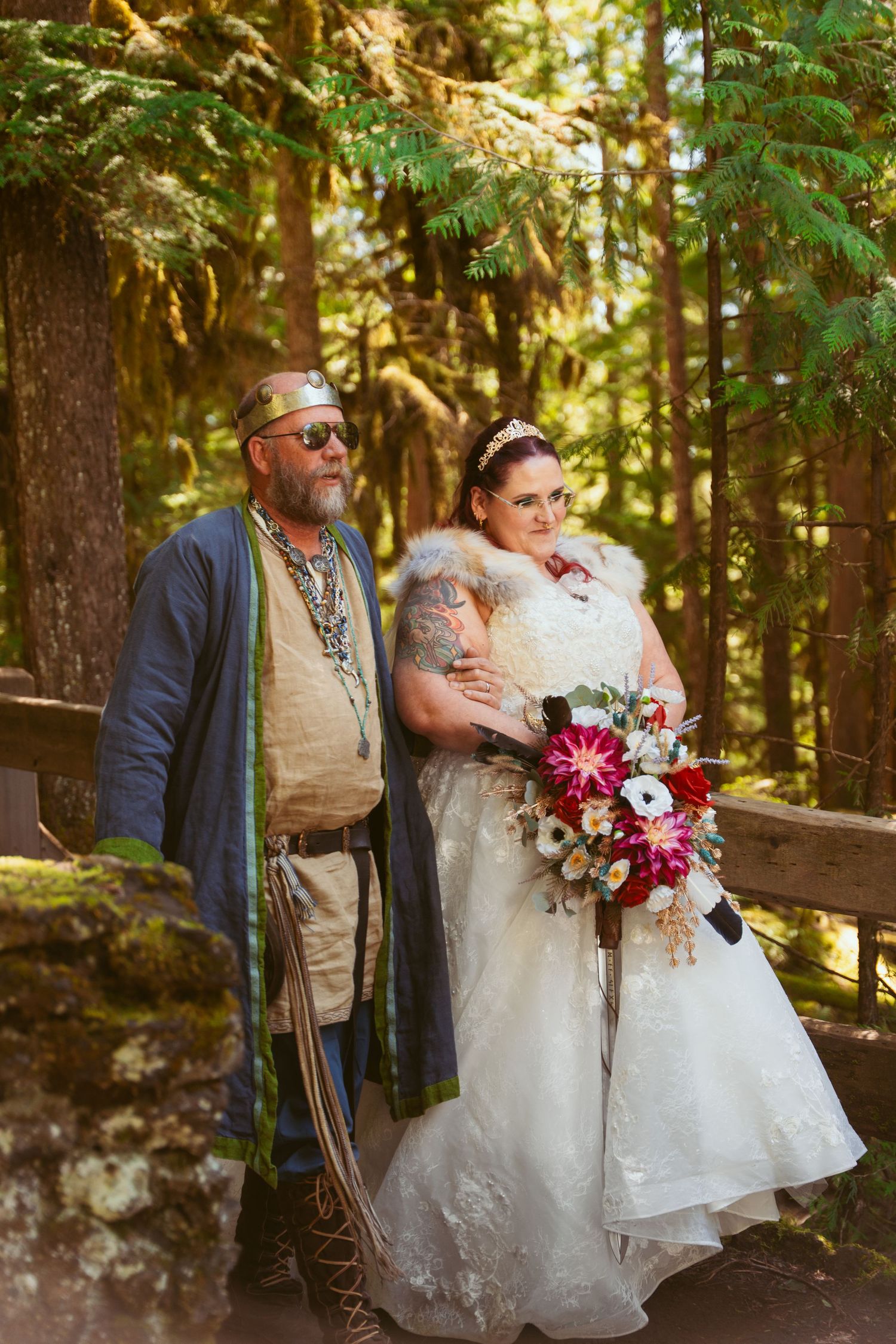 Couple in wedding attire embracing during outdoor forest ceremony with red floral bouquet.