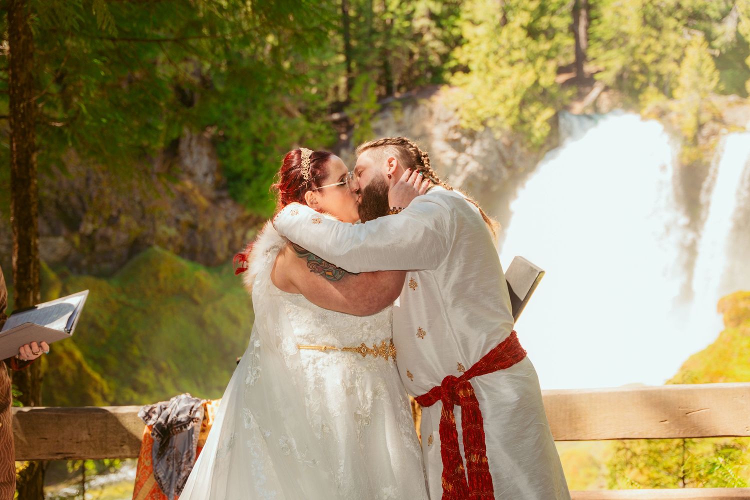 A wedding ceremony scene with a couple in white attire embracing by a waterfall.