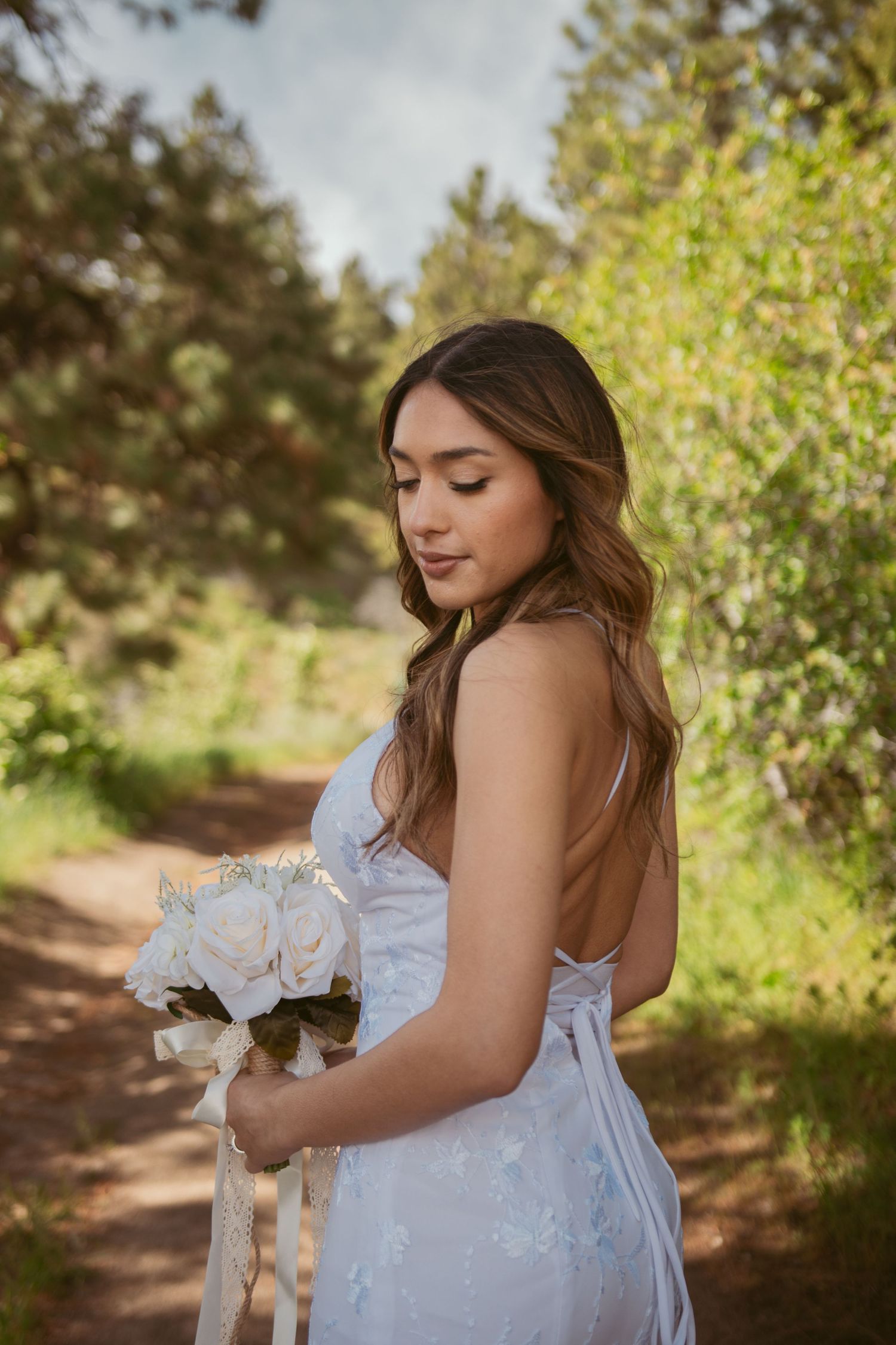 A bride in a white lace dress holds a bouquet of white roses on a nature trail.