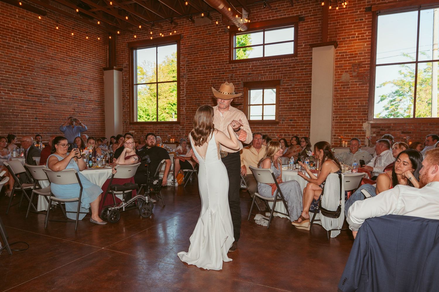 Dancers moving across wooden floor in sunlit brick venue during wedding celebration.