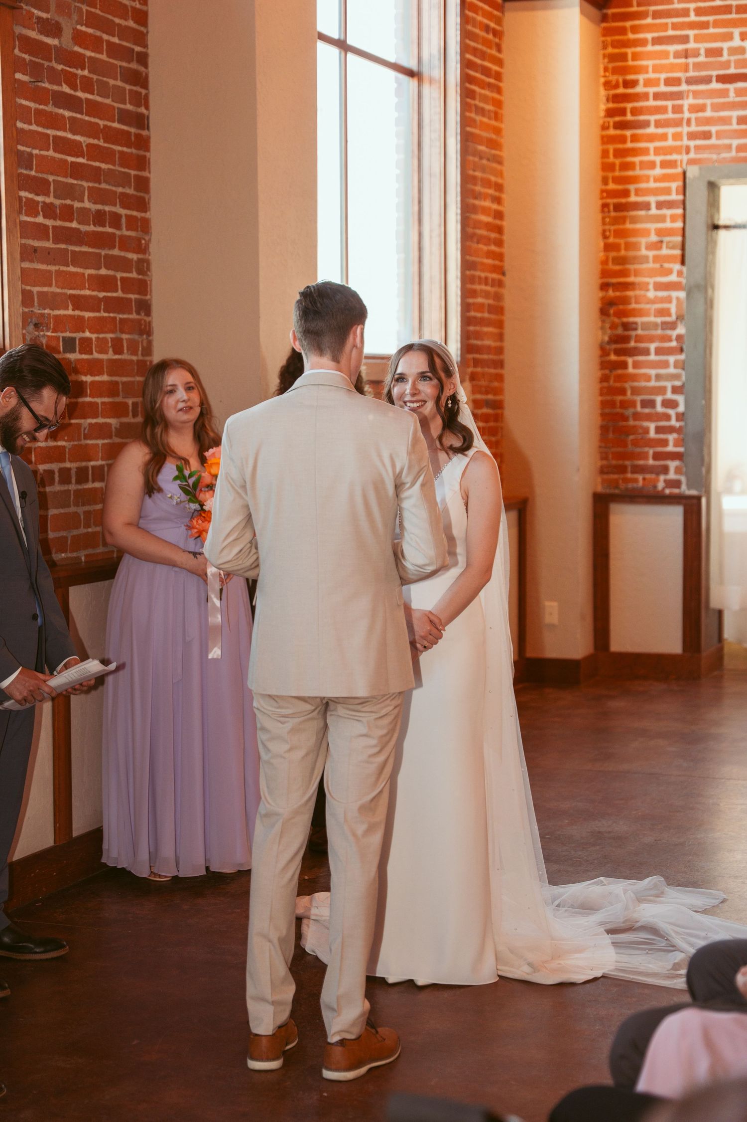 Wedding party gathered in sunlit brick venue with purple bridesmaid dresses.