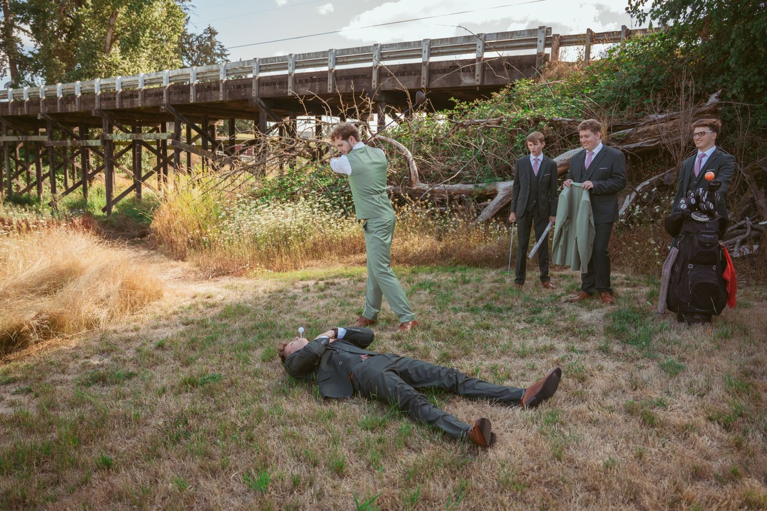 Wedding party members in green and gray attire pose progressively falling down on grassy slope.