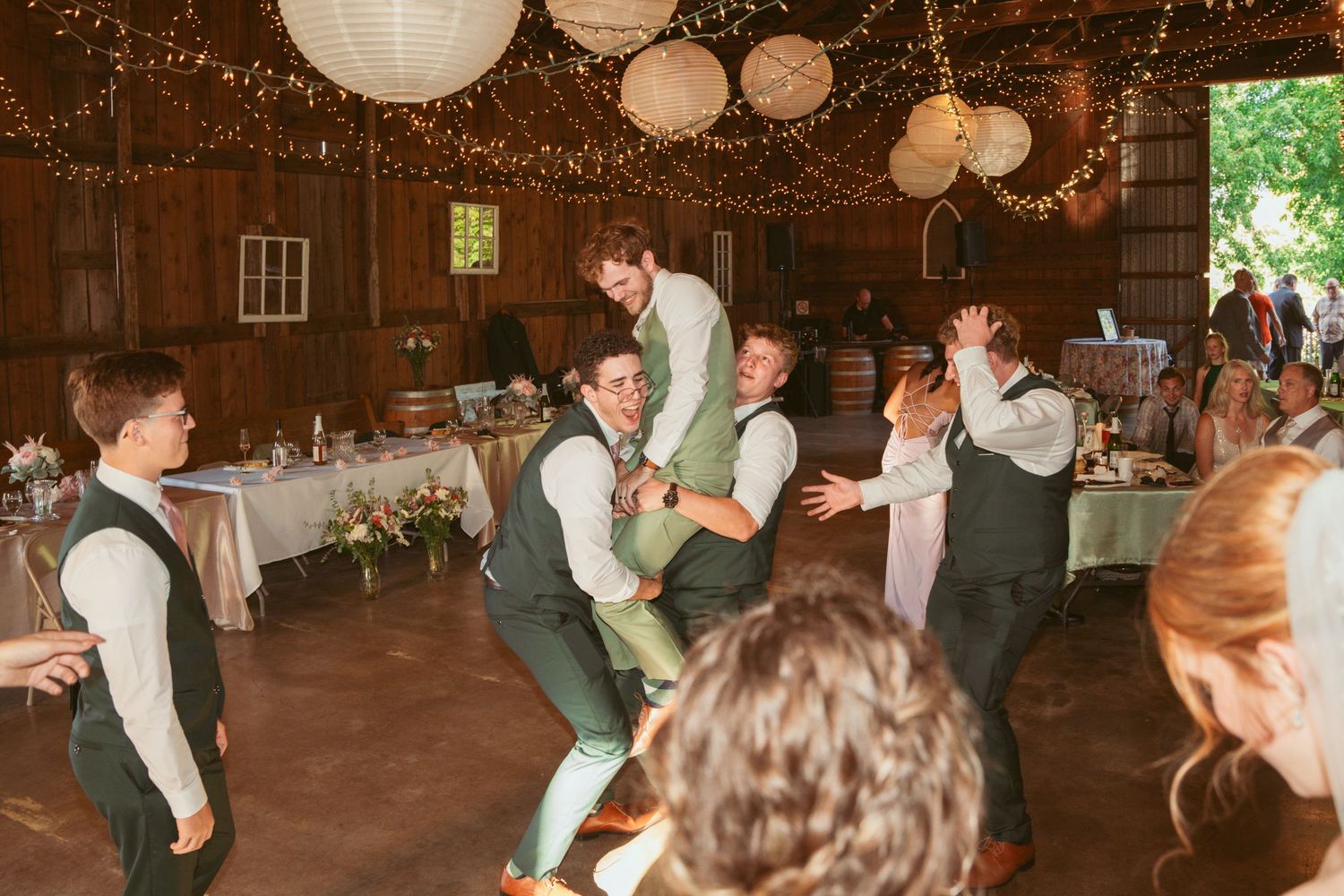 Wedding guests dancing energetically in a rustic barn venue decorated with string lights and paper lanterns.