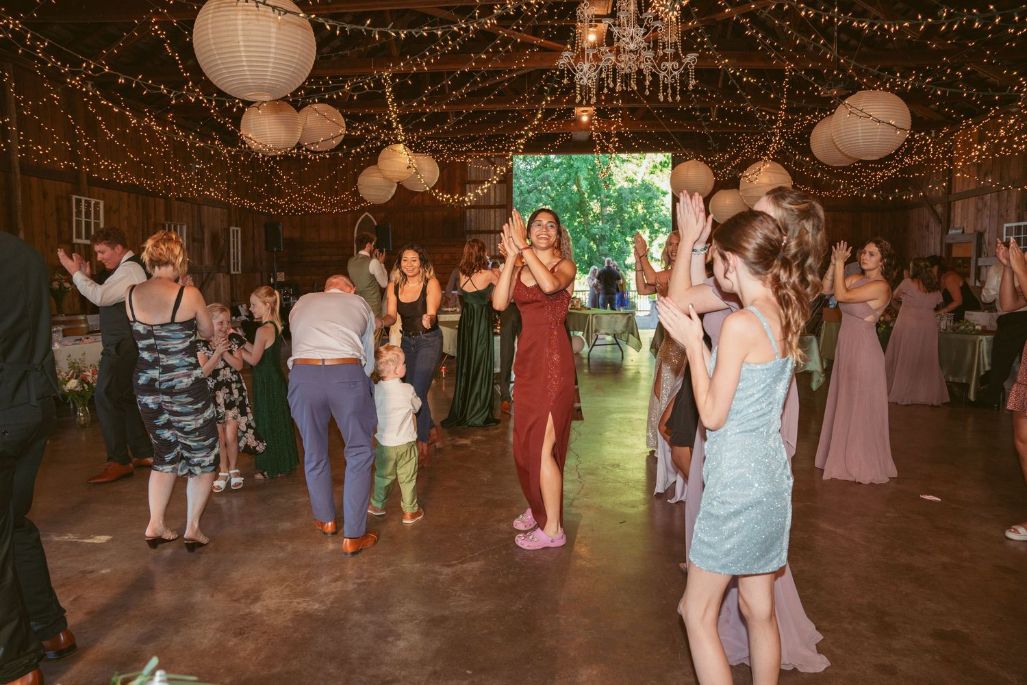 Wedding party dancing and celebrating in a barn decorated with twinkling lights and paper lanterns.