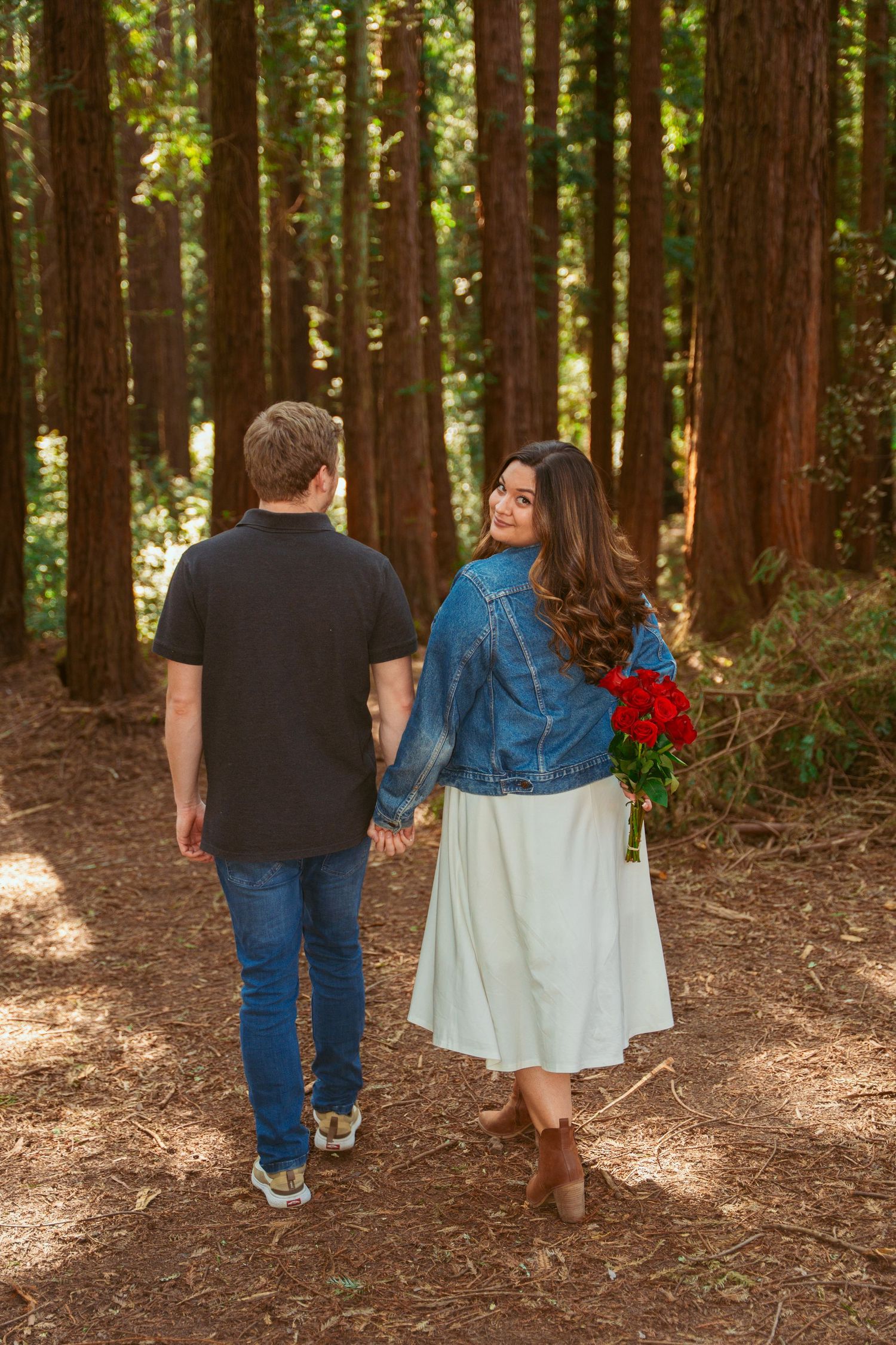 Couple walking hand in hand through redwood forest carrying red roses.