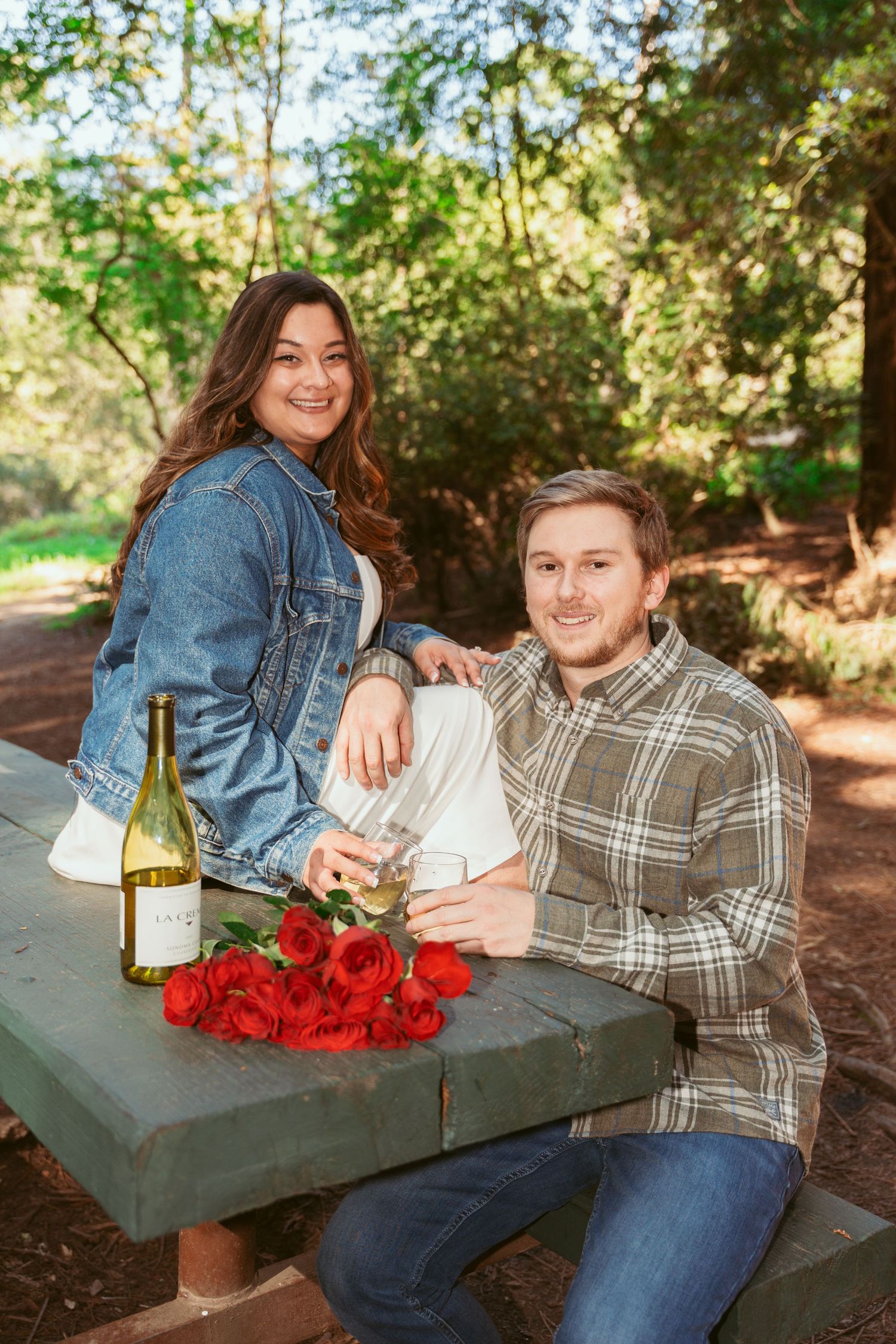 A couple sits together on a bench with red roses and wine in a peaceful forest setting.
