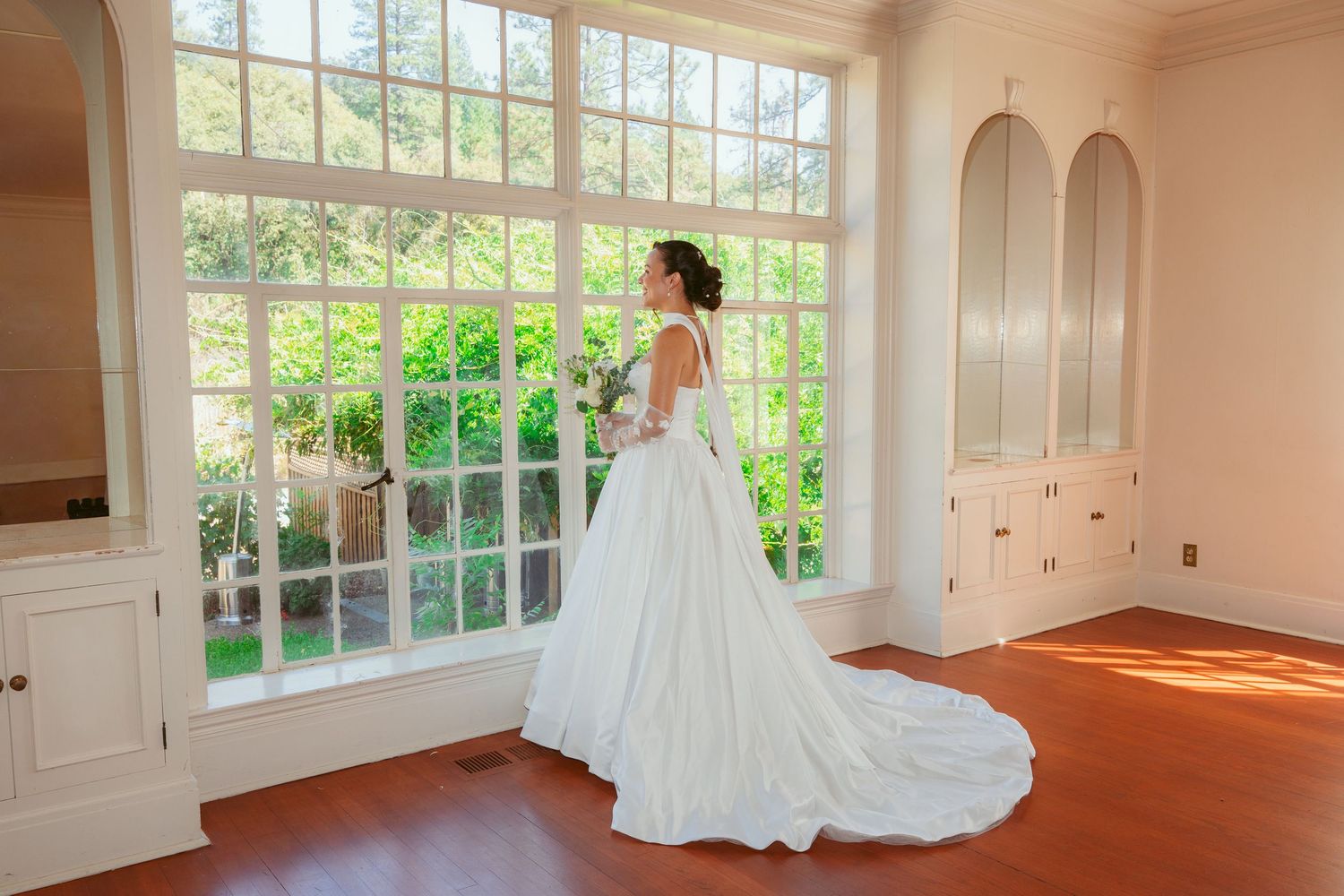 A bride in a flowing white wedding gown stands by a large window in an elegant room with natural light.