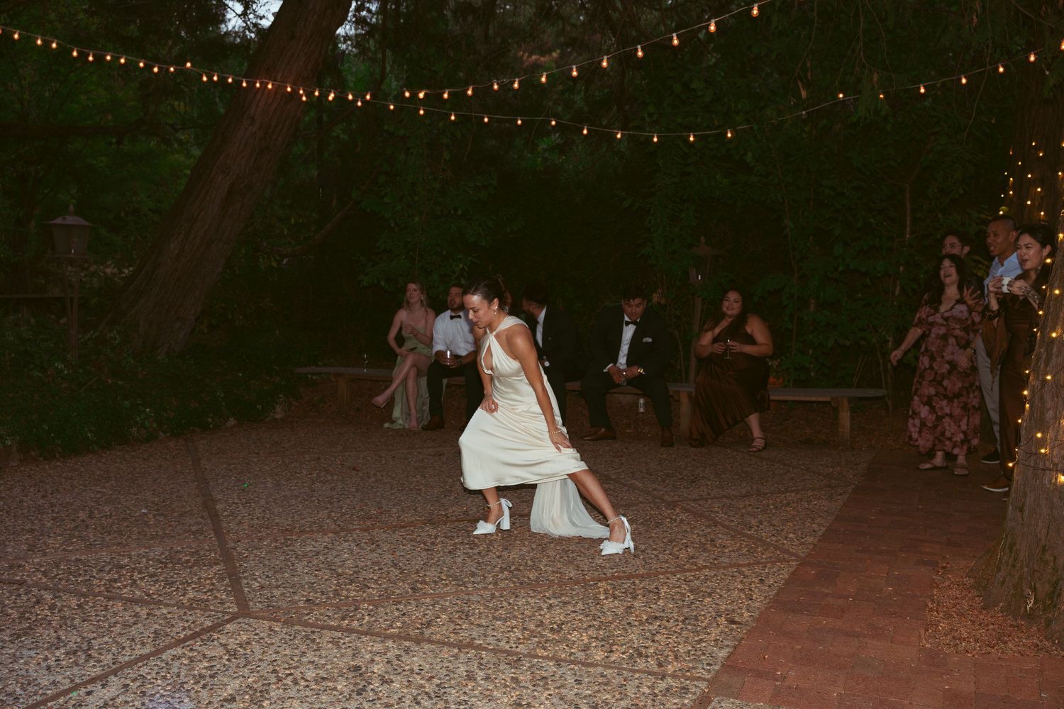 Guests dance and celebrate at an outdoor wedding reception under twinkling string lights at dusk.