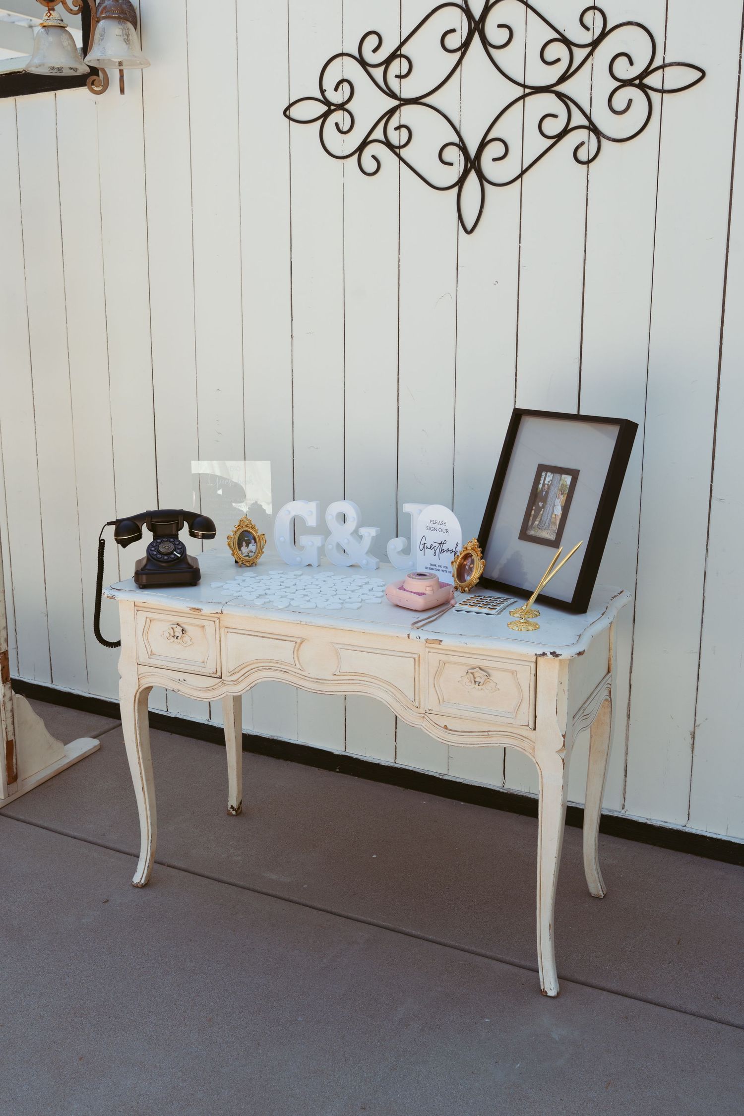 Vintage-style white table with ornate wall decor displaying wedding items and frames.