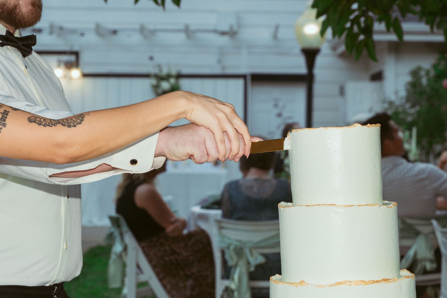 Close-up shot of hands cutting into a minimalist three-tiered white wedding cake outdoors.