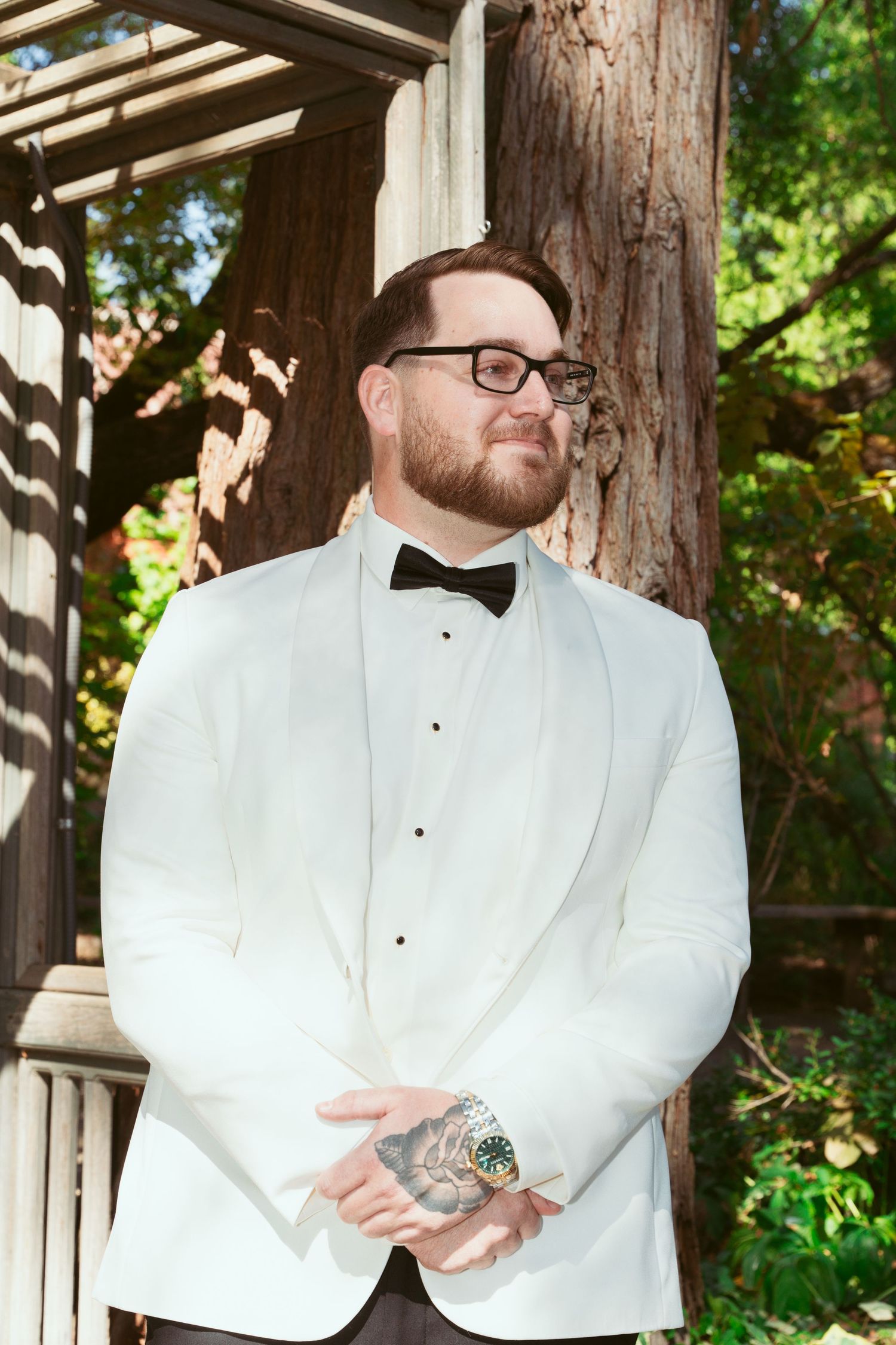 Groom in white tuxedo jacket and bow tie poses thoughtfully under wooden gazebo.