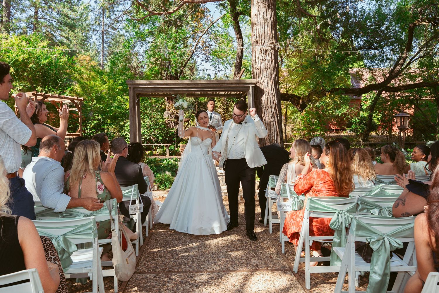 Wedding guests in mint green dresses gather outdoors under tall trees.