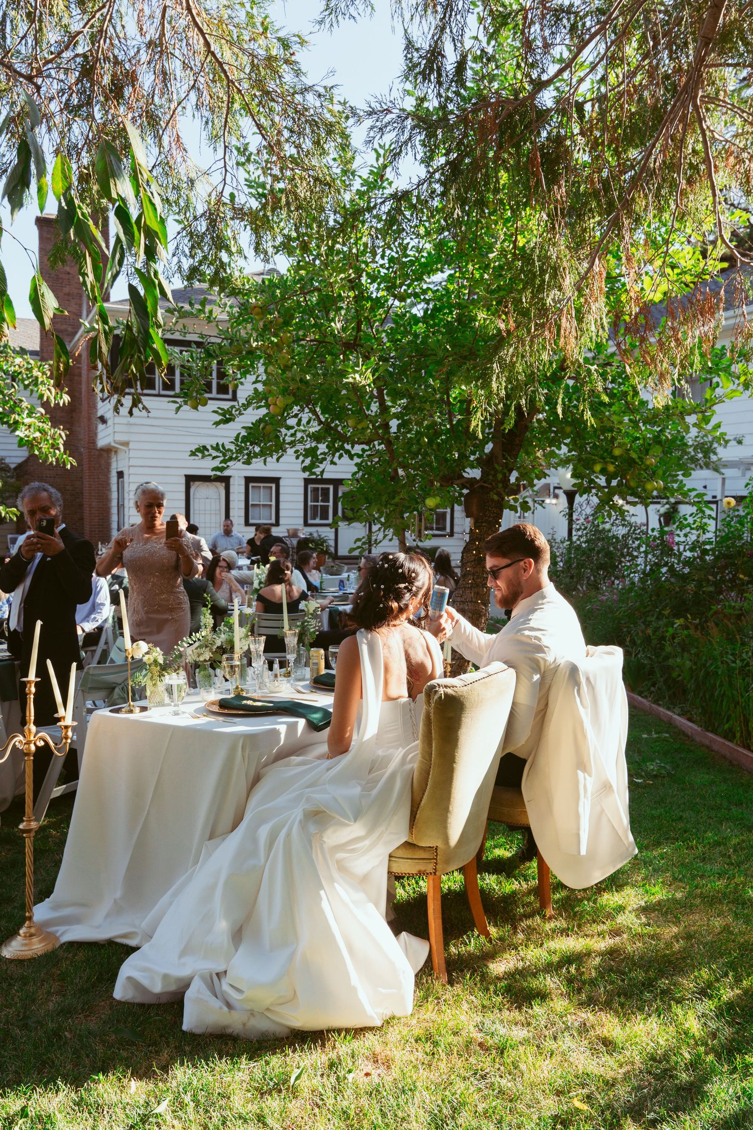 Wedding guests enjoy an outdoor reception with white-clothed tables under trees in a garden setting.