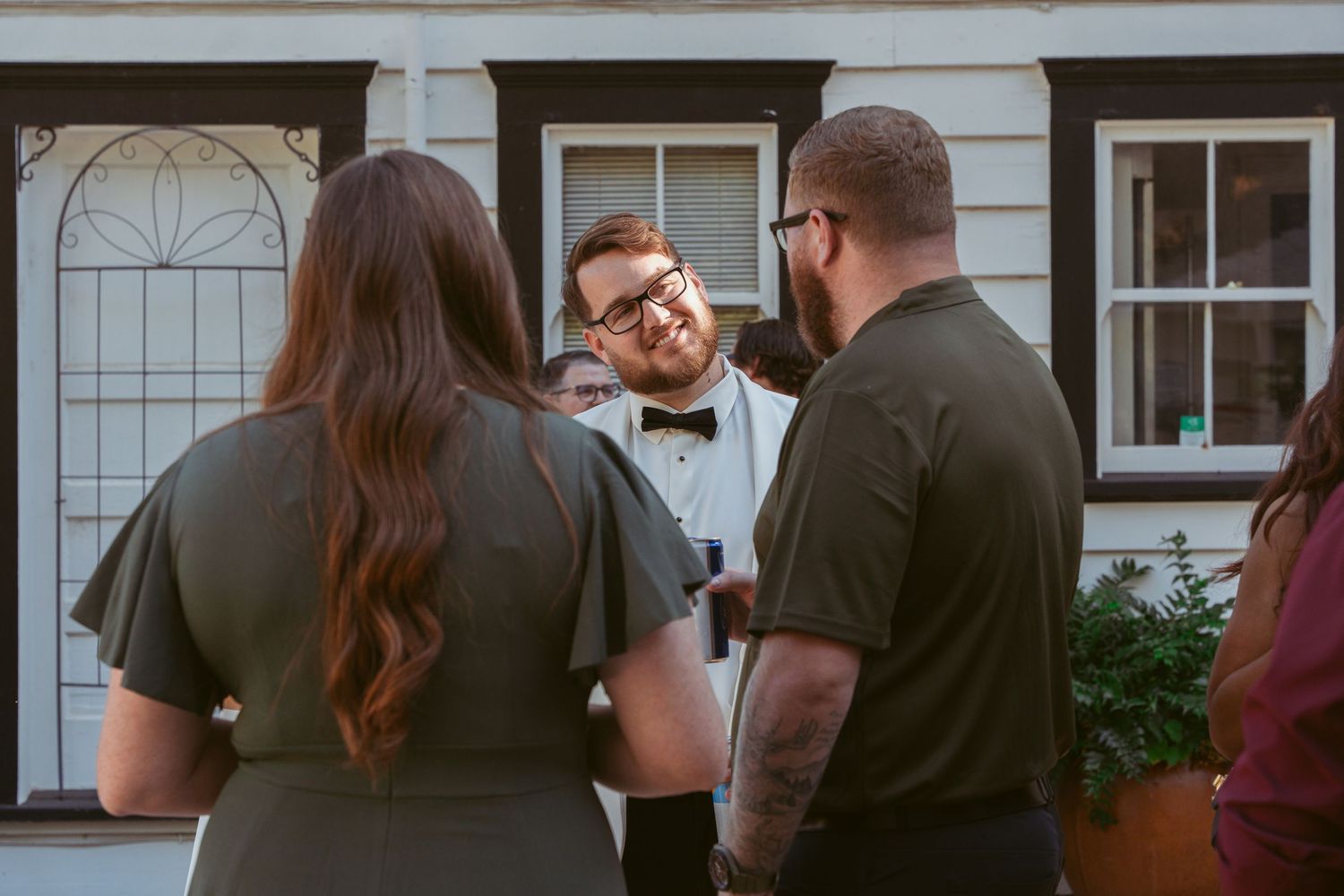 Wedding party members in formal attire stand together against a white building exterior.