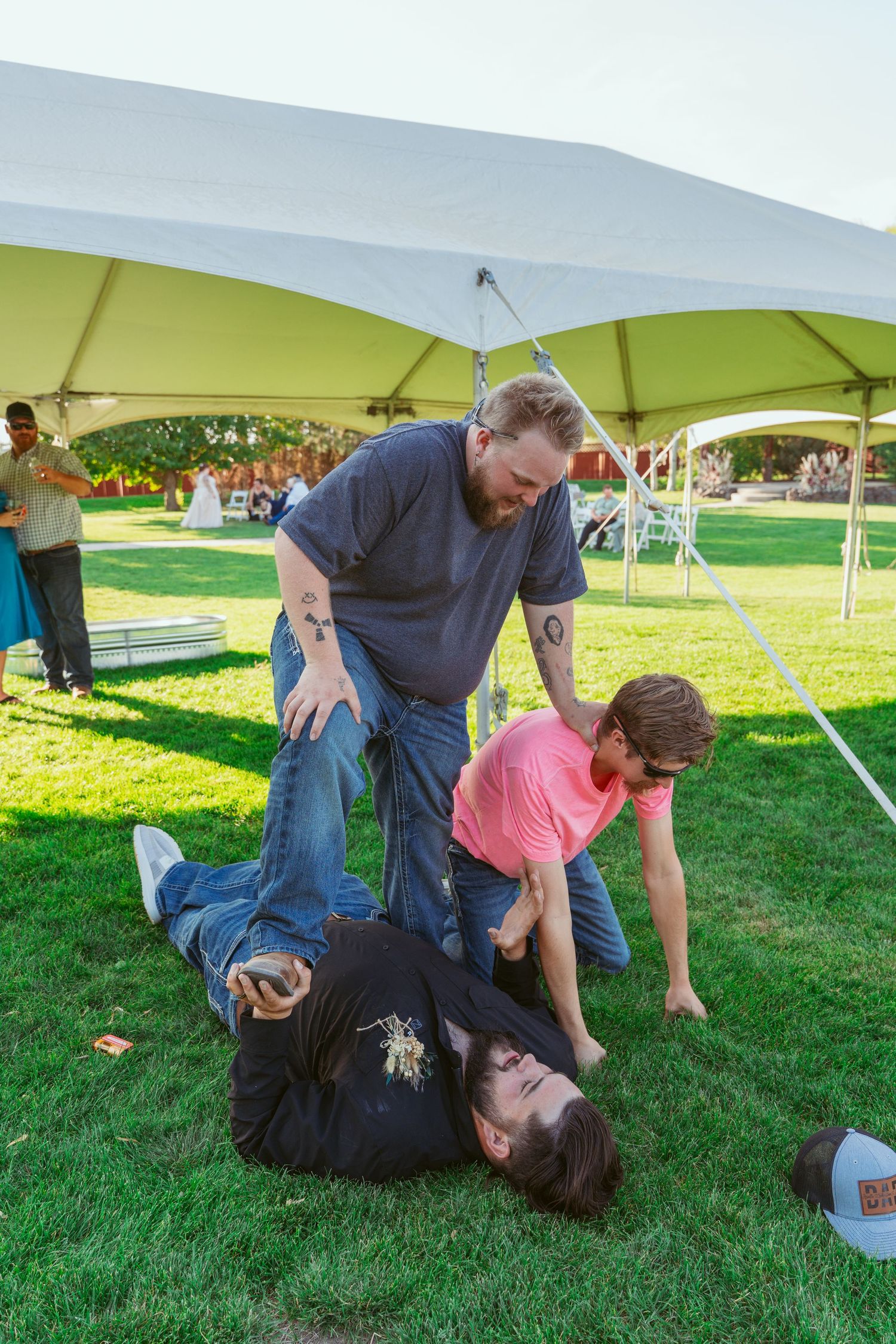 Playful wedding guests wrestling on grass under white tents during outdoor reception.