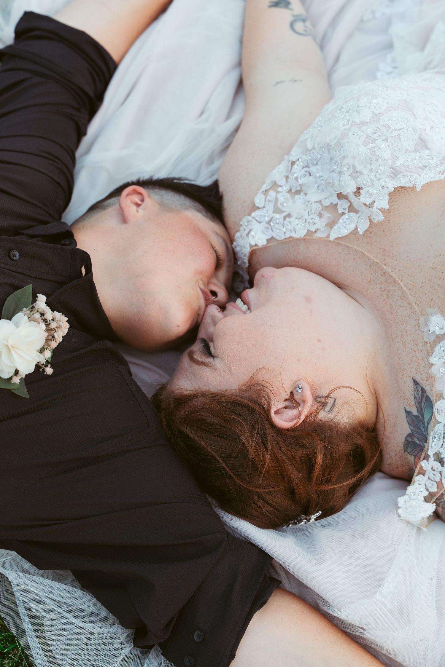 A couple in wedding attire lying on grass together with their faces close.