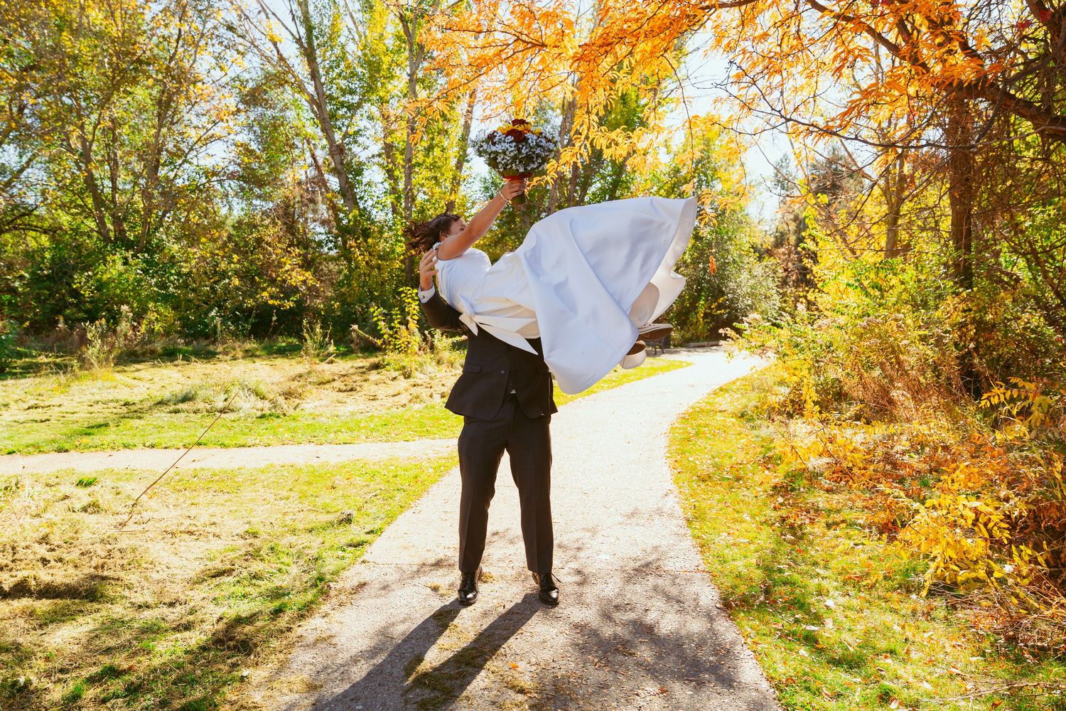 A wedding couple sharing a playful moment on a path with autumn trees in the background.