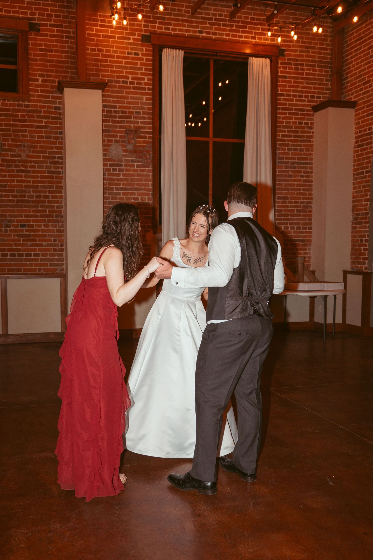 Wedding guests dancing in formal attire under string lights in a brick-walled reception space.