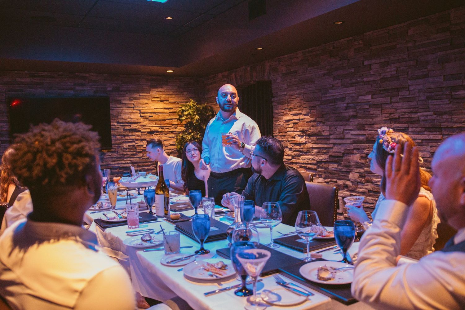 Wedding reception guests gather around tables in a dimly lit restaurant with stone walls.
