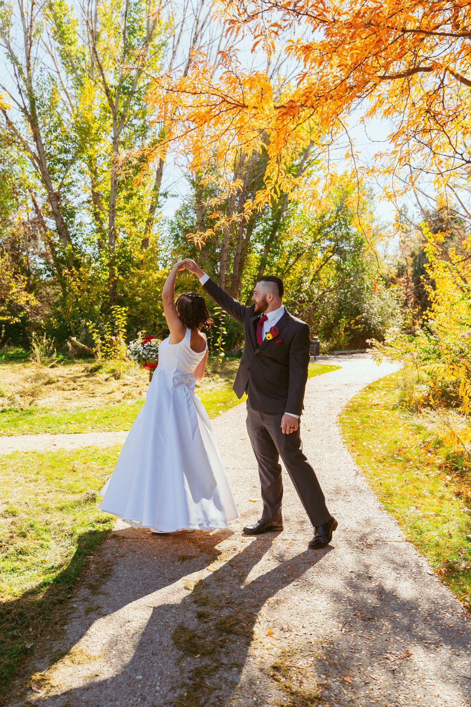 Playful wedding couple dancing on dirt path surrounded by golden autumn trees.