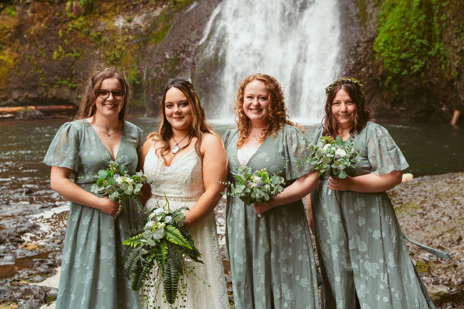 Joyful wedding party poses for photos in sage green dresses by a waterfall.
