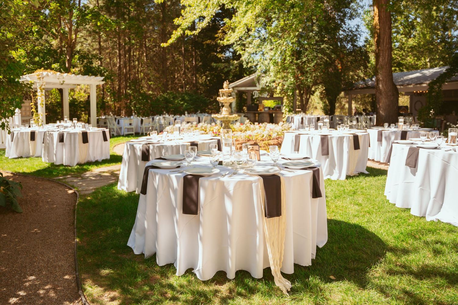 Elegant outdoor wedding reception setup with white-clothed tables arranged under tall trees in a garden setting.