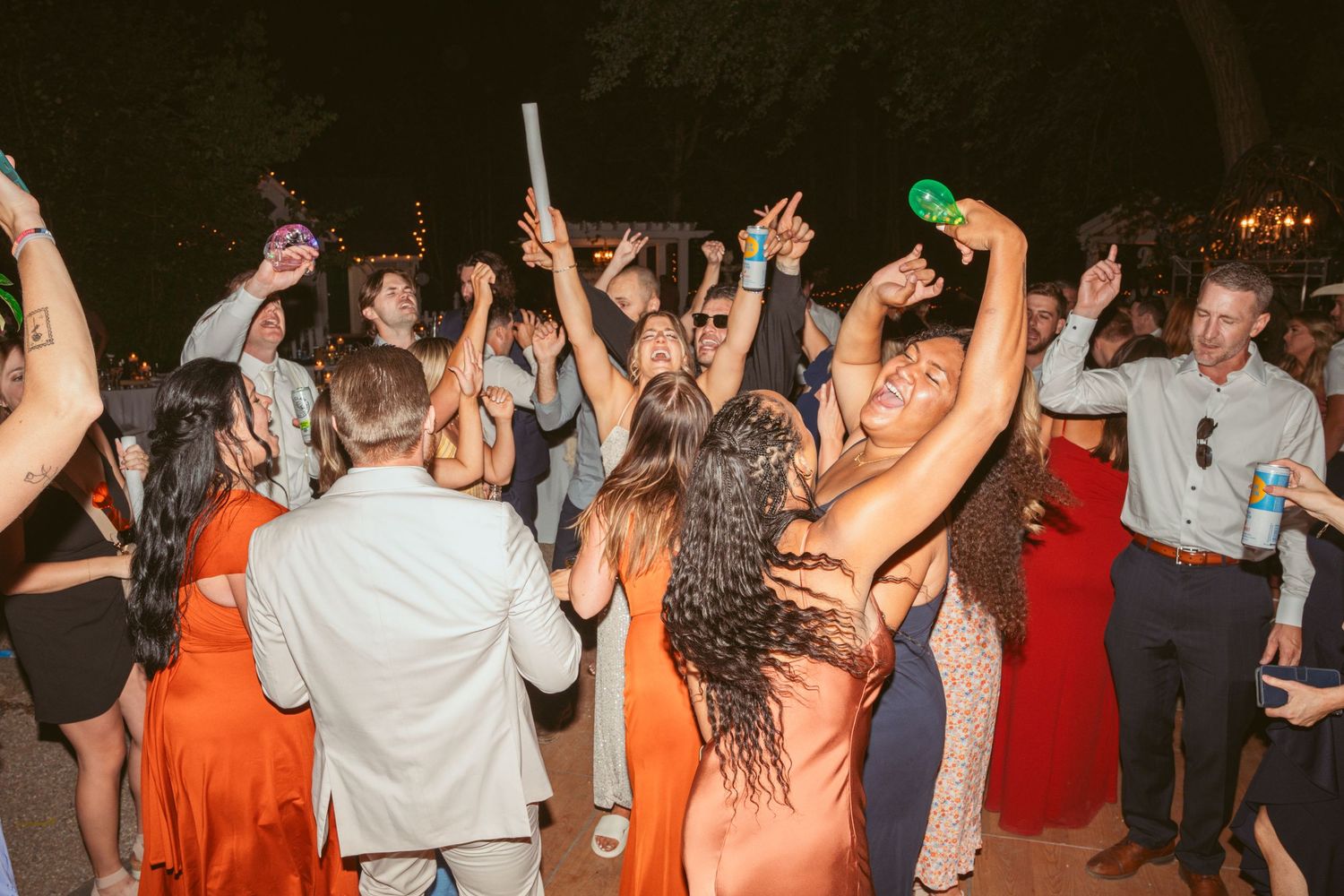 Energetic crowd dancing and celebrating with raised drinks at an outdoor evening party.