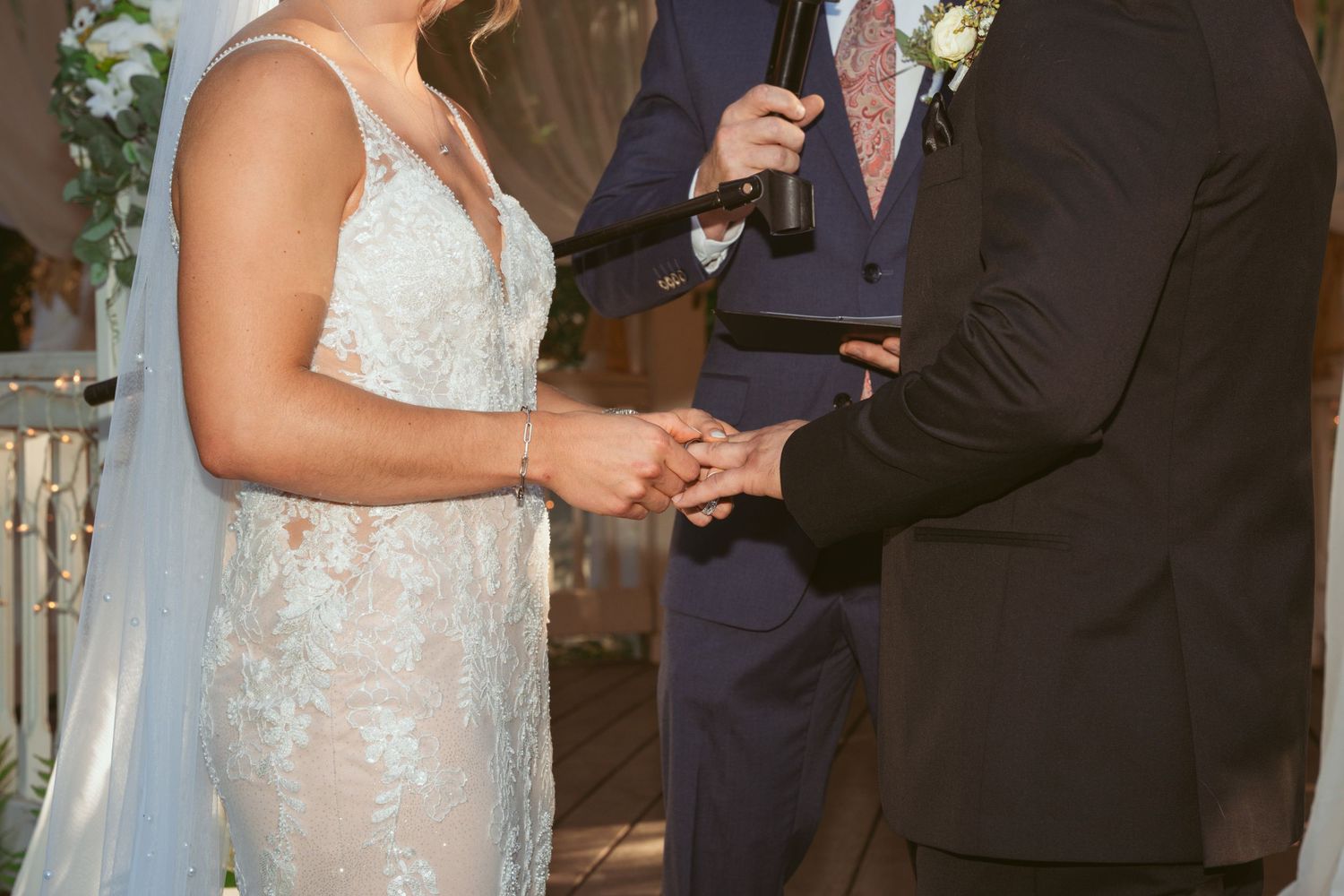Close-up of wedding couple holding hands during ring exchange ceremony in an outdoor setting.