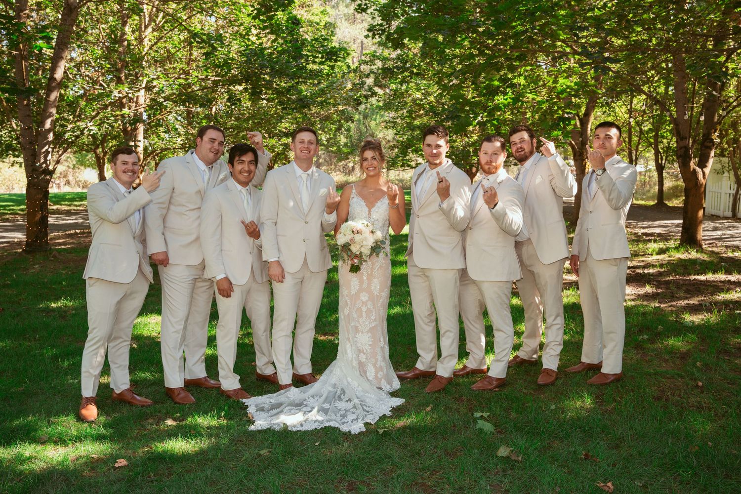 Wedding party in cream colored attire poses together on a sunny lawn surrounded by trees.