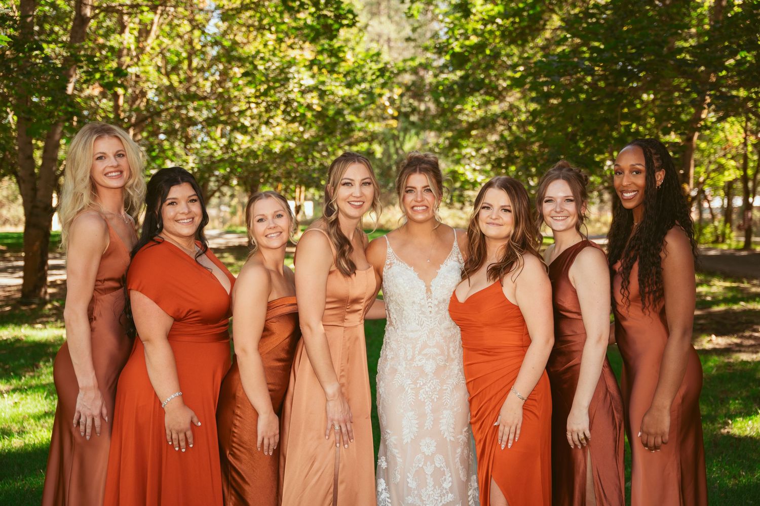 Wedding party in coordinated orange and white dresses poses together on a grassy lawn under trees.