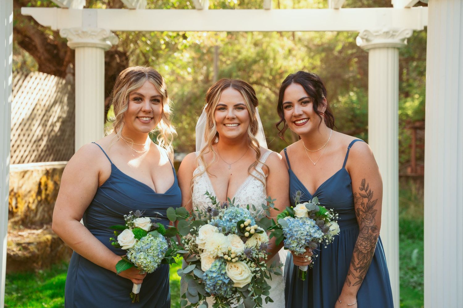 Bridesmaids in navy dresses hold blue and white bouquets while standing under a white arbor.