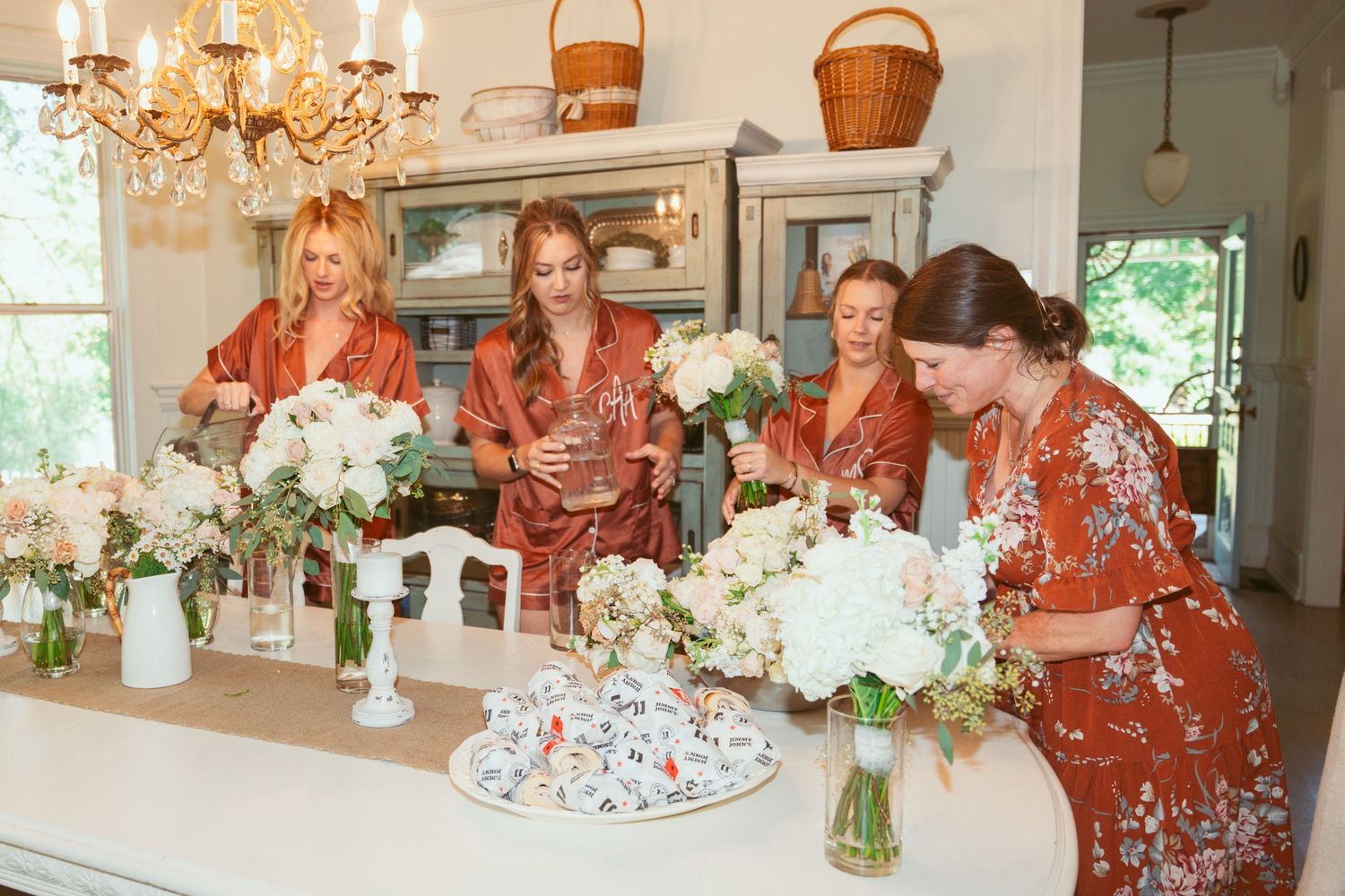 A panoramic view of a bridal party in matching rust-colored robes gathered in an elegant room with chandeliers.
