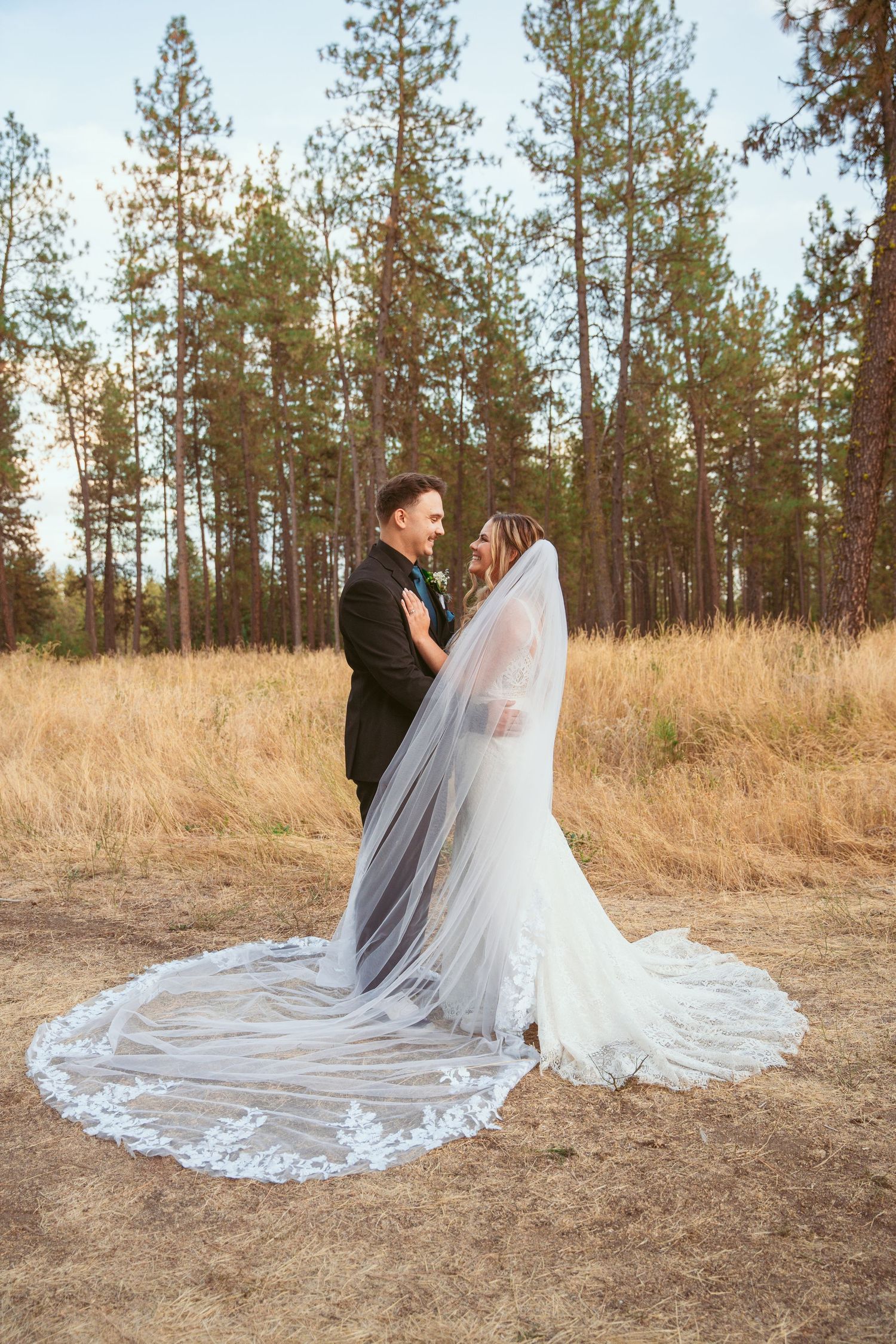 A wedding couple poses in a field of tall grass with pine trees in the background as the bride's veil flows dramatically.
