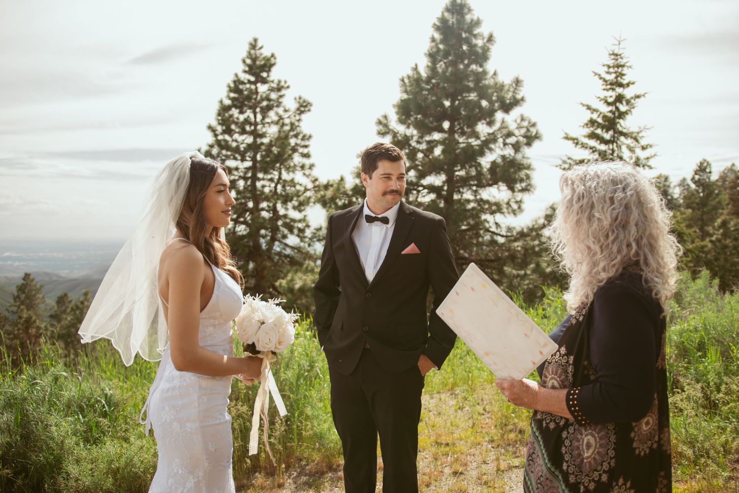 An outdoor wedding ceremony takes place on a mountain trail with evergreen trees and valley views.