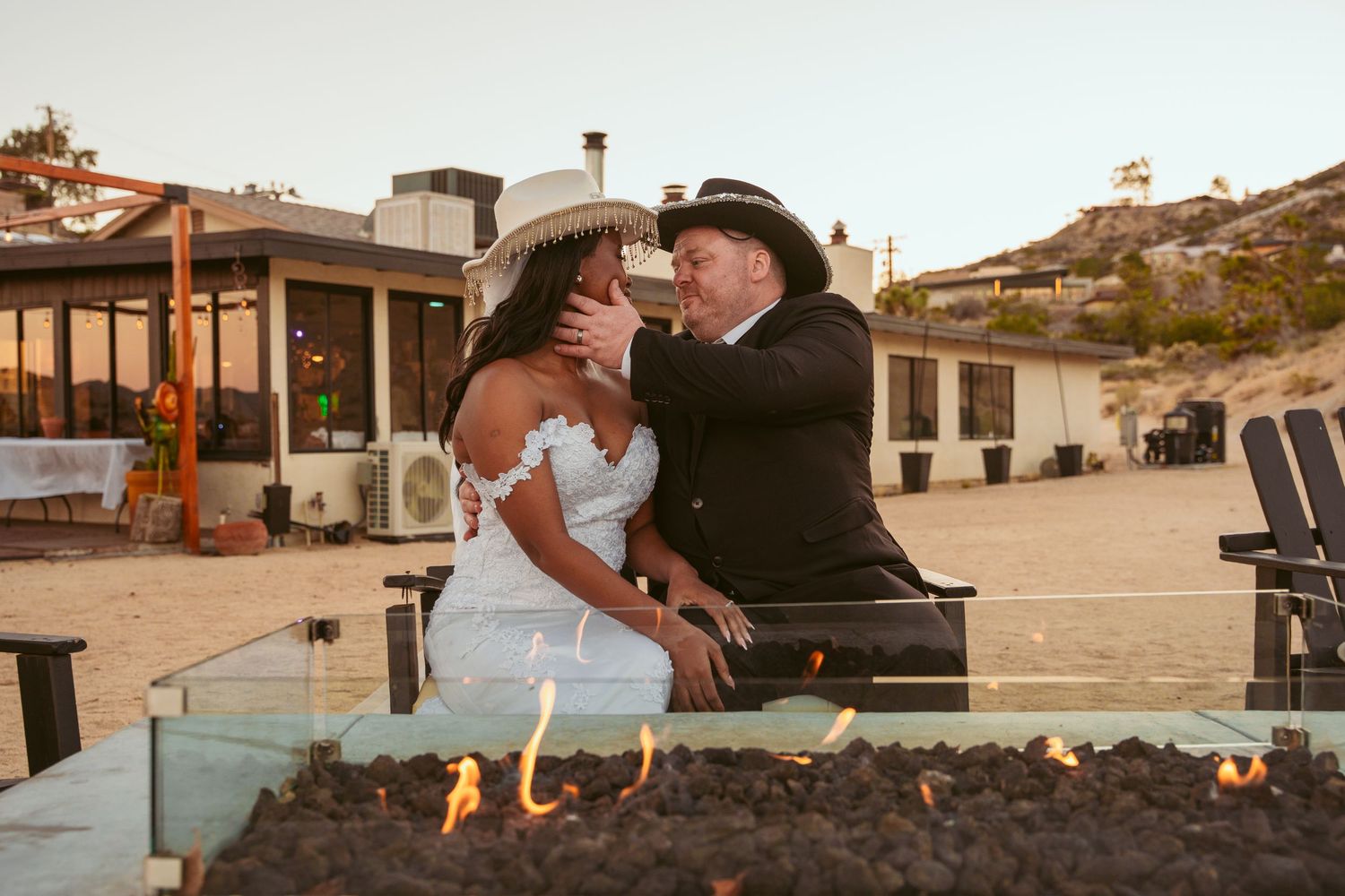 Newlyweds sit together by an outdoor fire pit at sunset.