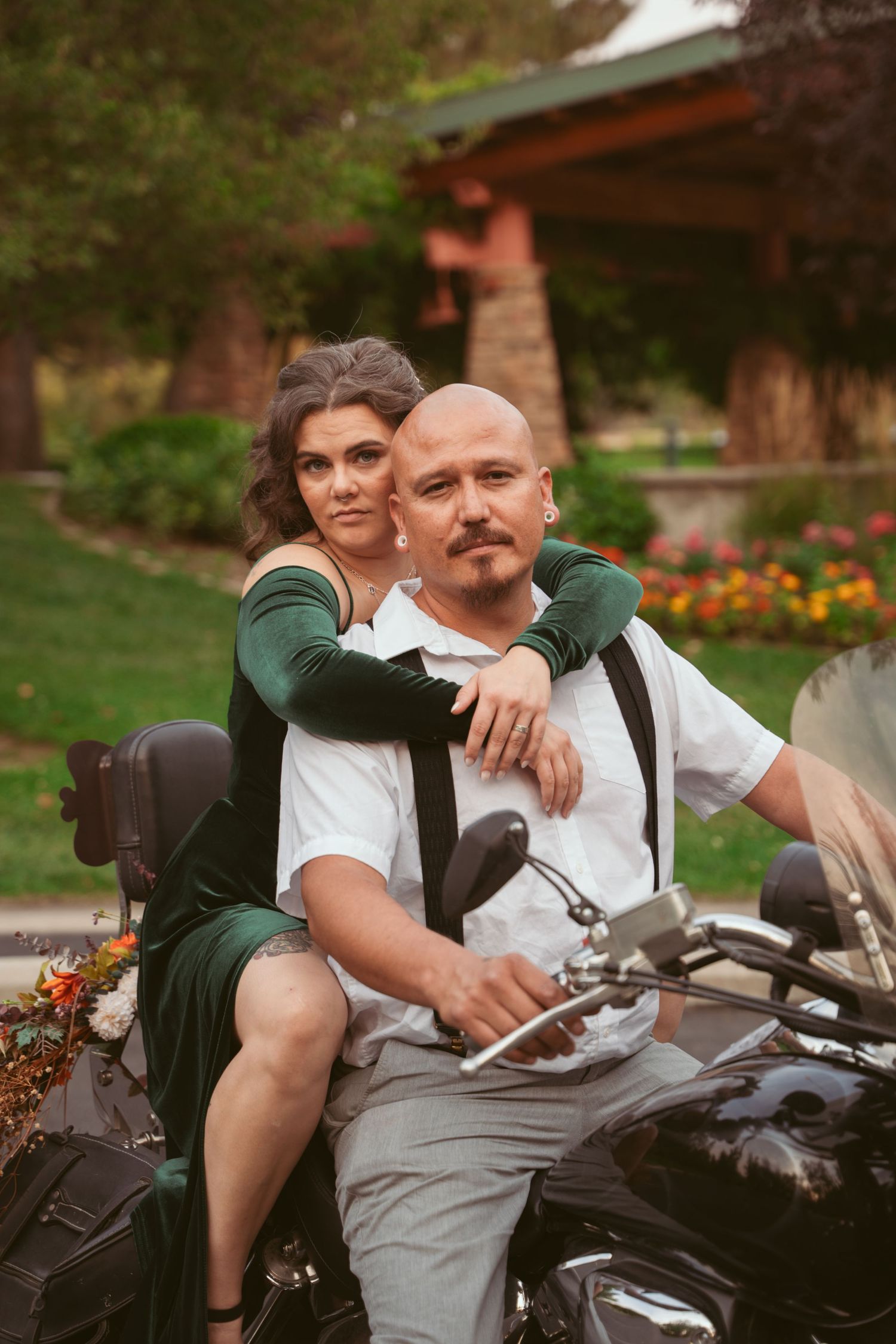 Couple posing on a motorcycle with flowers and greenery in the background.