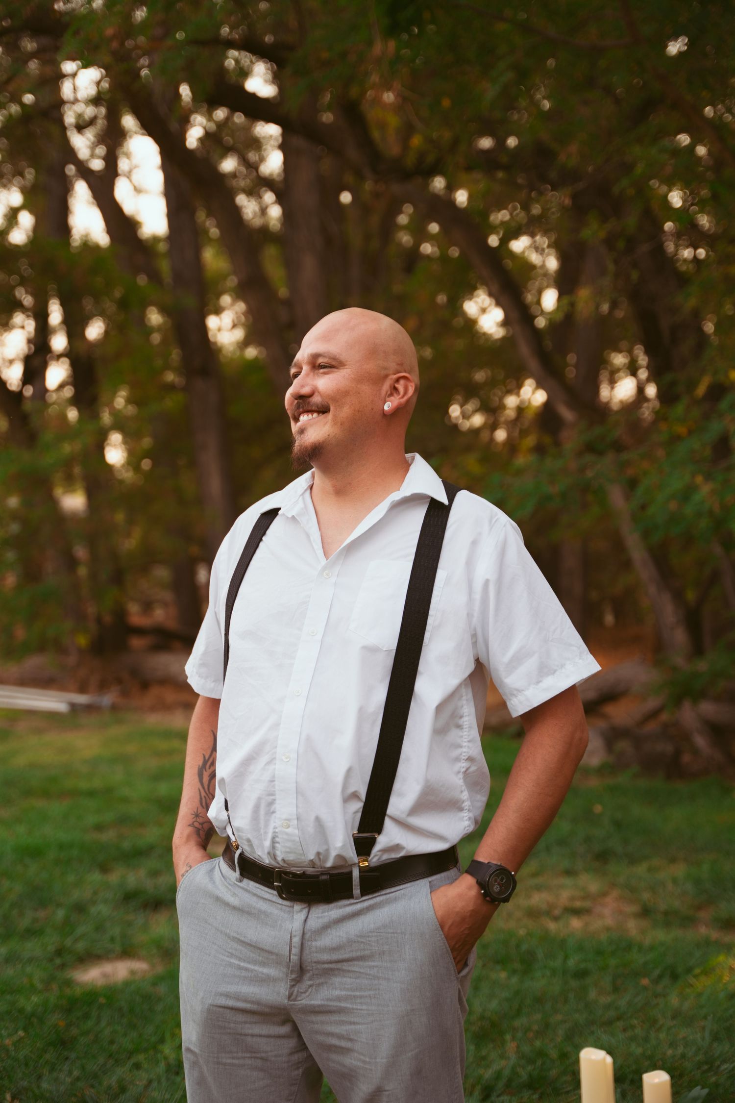 Groom in white shirt and suspenders poses outdoors during golden hour wedding photoshoot.