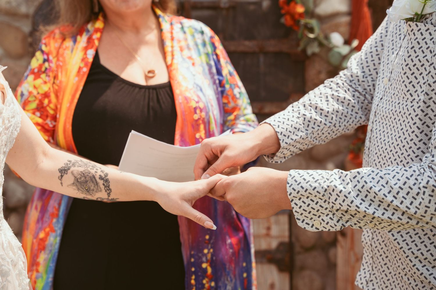 Close-up of hands exchanging rings during a wedding ceremony with colorful floral backdrop.