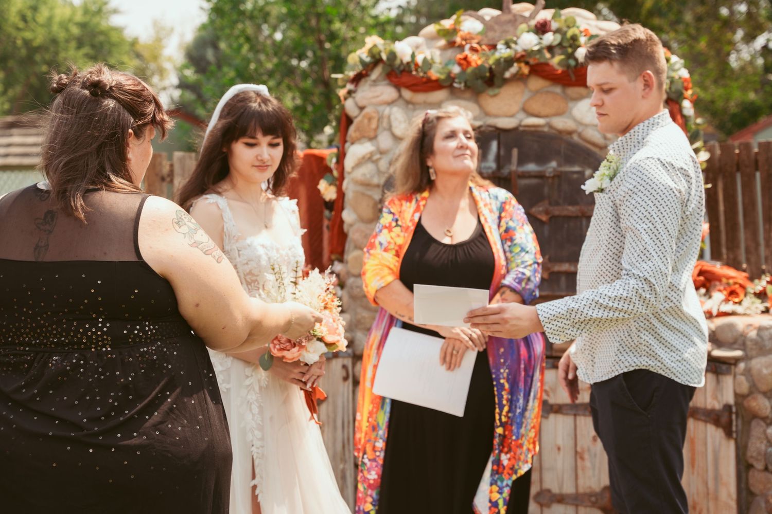 Wedding ceremony participants stand in a line outdoors with decorative stone backdrop.