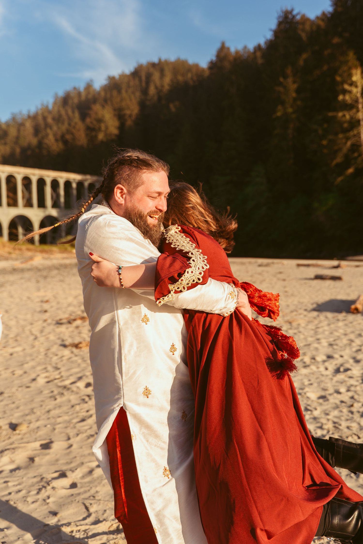 A couple laughs together during a romantic beach photoshoot with cliffs and an arch bridge in the distance.