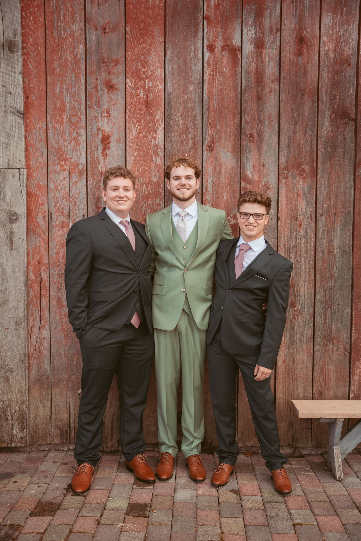 Groomsmen pose together in black and sage green suits against weathered wooden barn background.