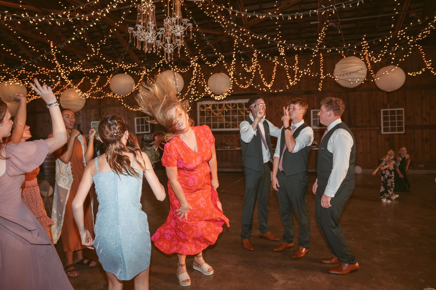 People dancing in a rustic barn decorated with string lights and paper lanterns.