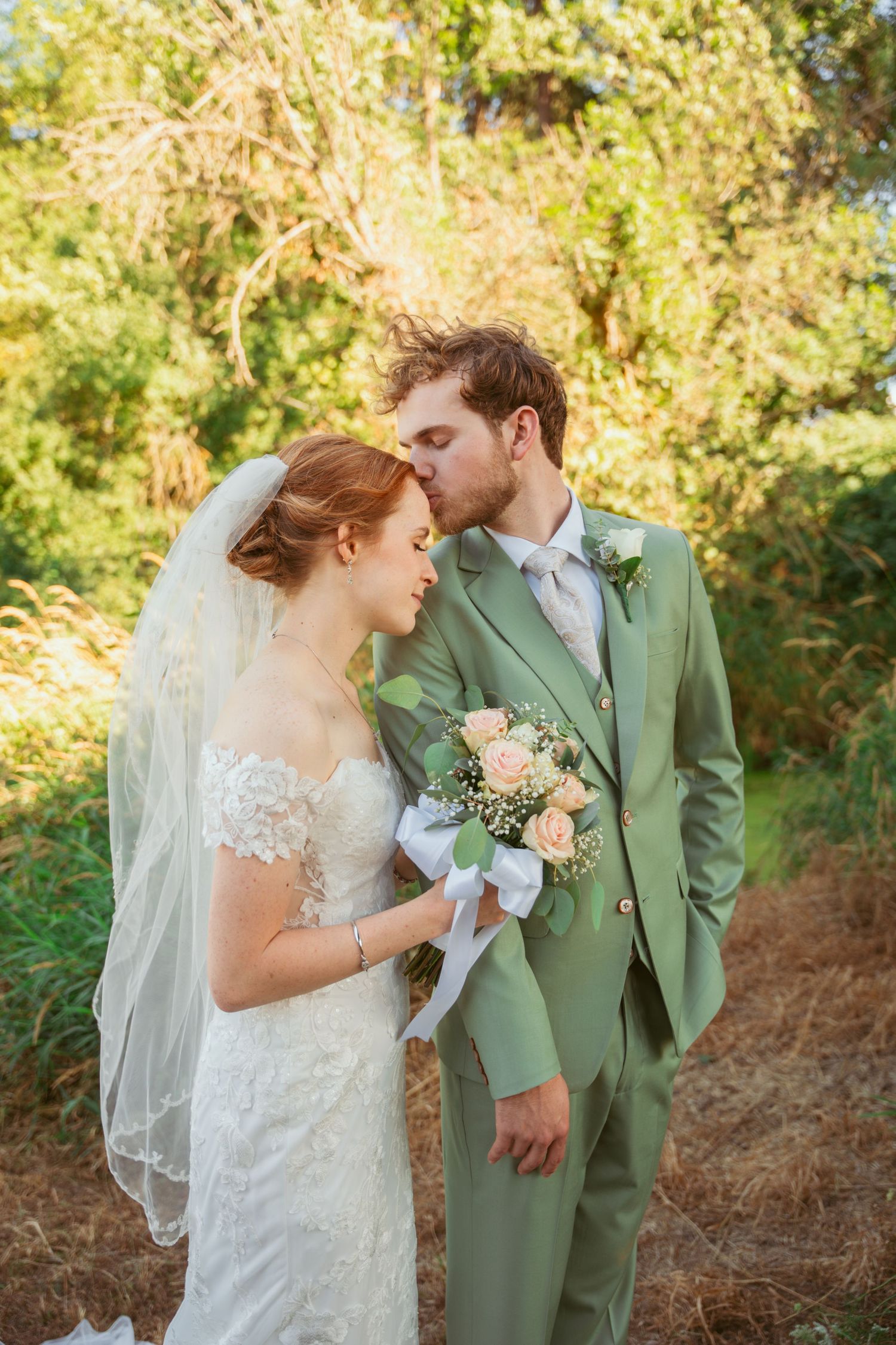 Romantic wedding couple in sage green and white share intimate moments against sunlit foliage.