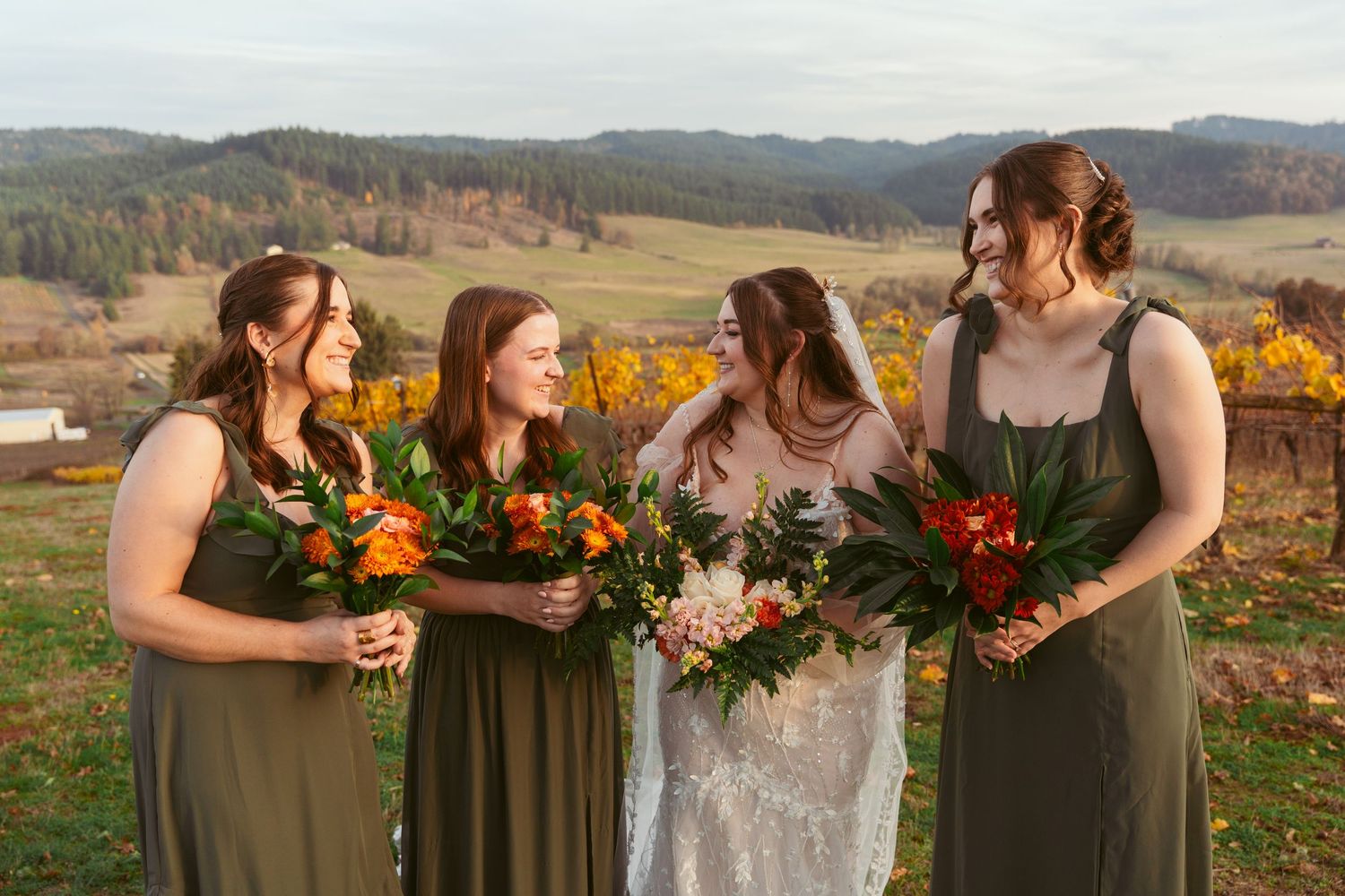 Bridesmaids in sage dresses share joyful moment with bride while holding vibrant fall bouquets.