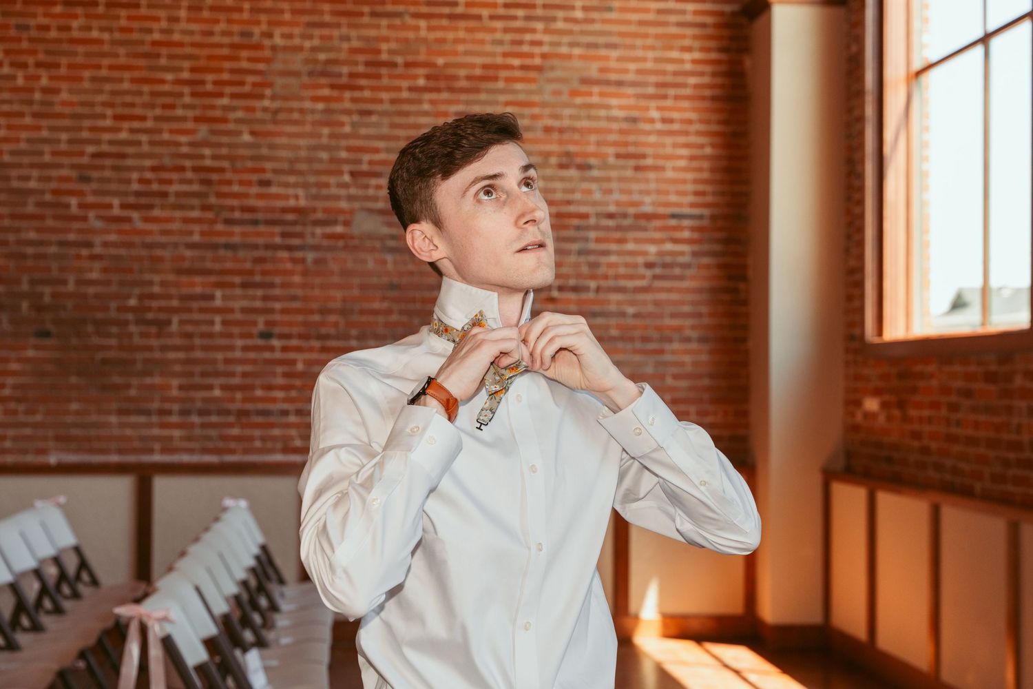 Person in white shirt adjusts tie while standing in sunlit room with brick walls.