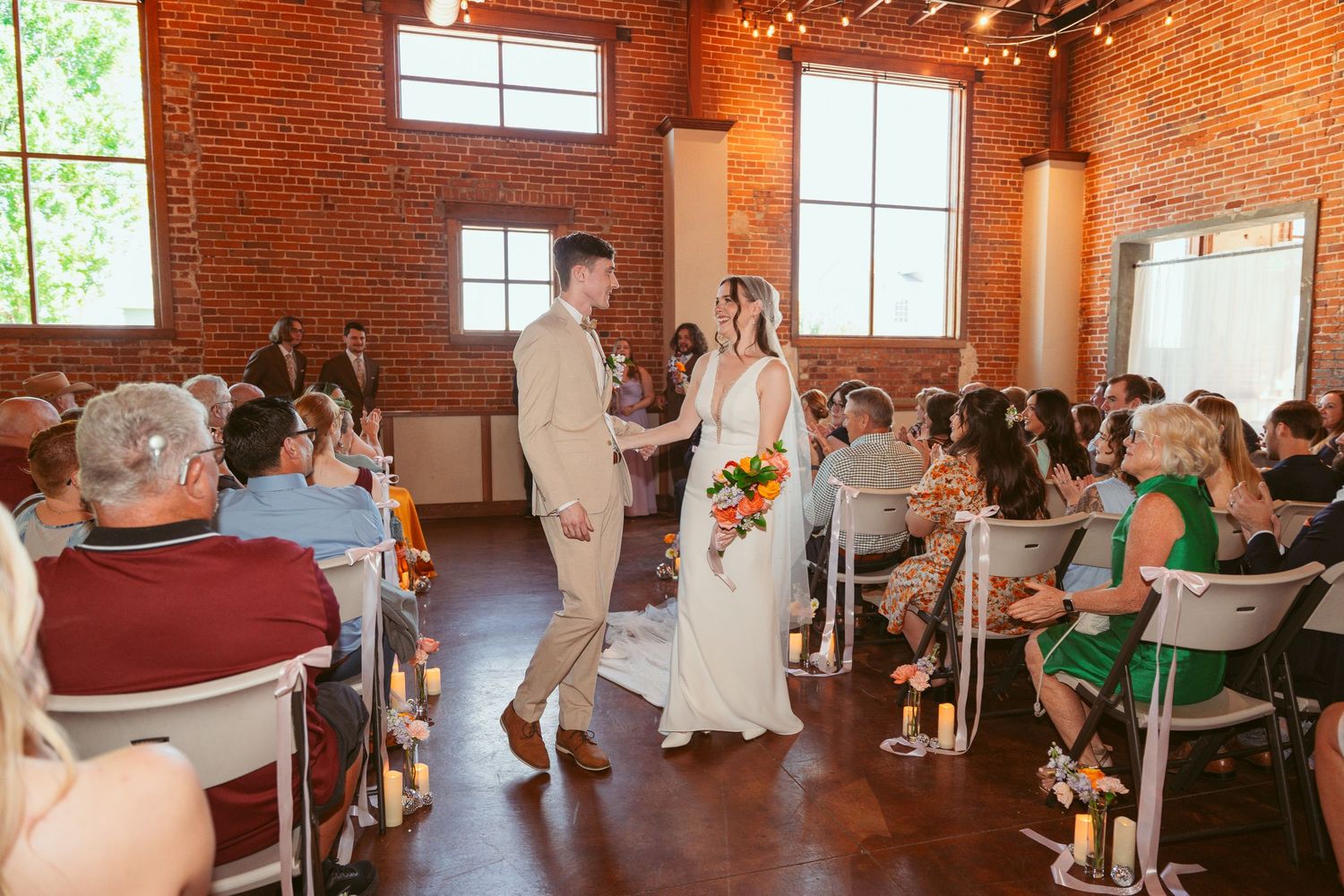 Wedding ceremony guests seated in a rustic brick venue with string lights and natural lighting from windows.