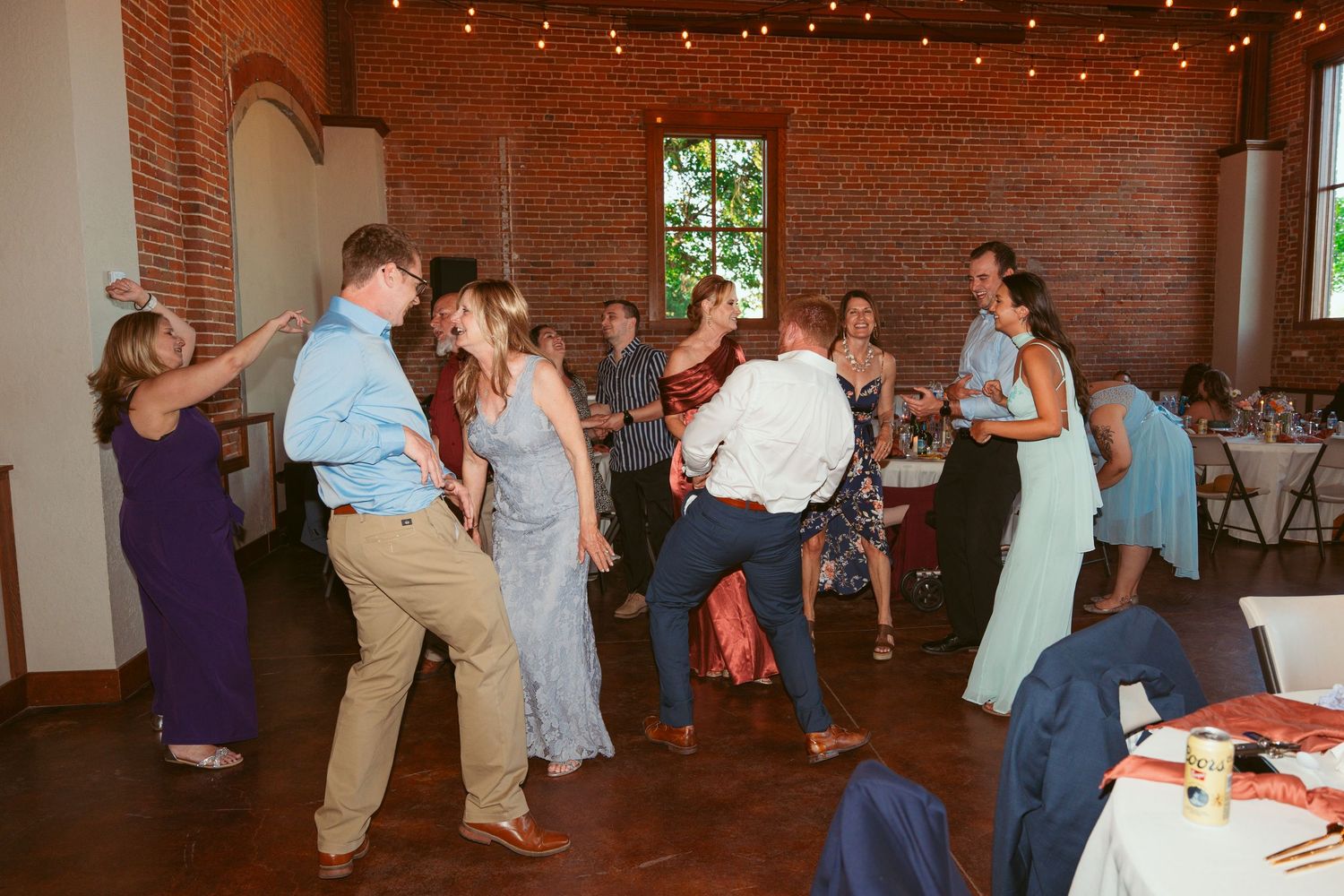 Lively wedding reception with guests dancing in groups on a spacious dance floor with brick walls.