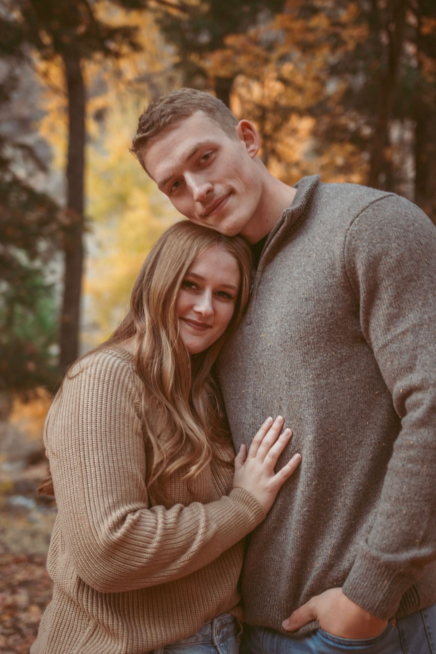 A couple embraces in an autumn forest setting with warm golden light filtering through trees.