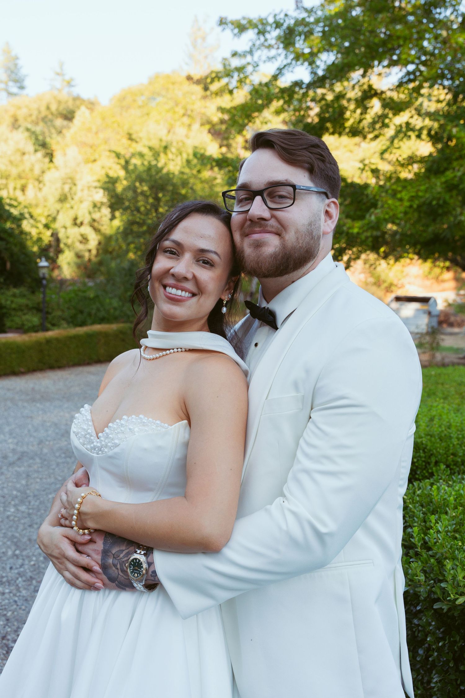 A joyful wedding couple embracing outdoors in formal attire against a sunlit garden backdrop.