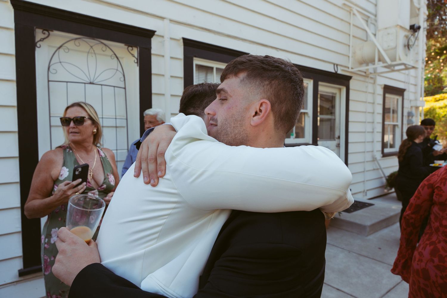 Wedding celebration with guests embracing on an outdoor patio near a white building.